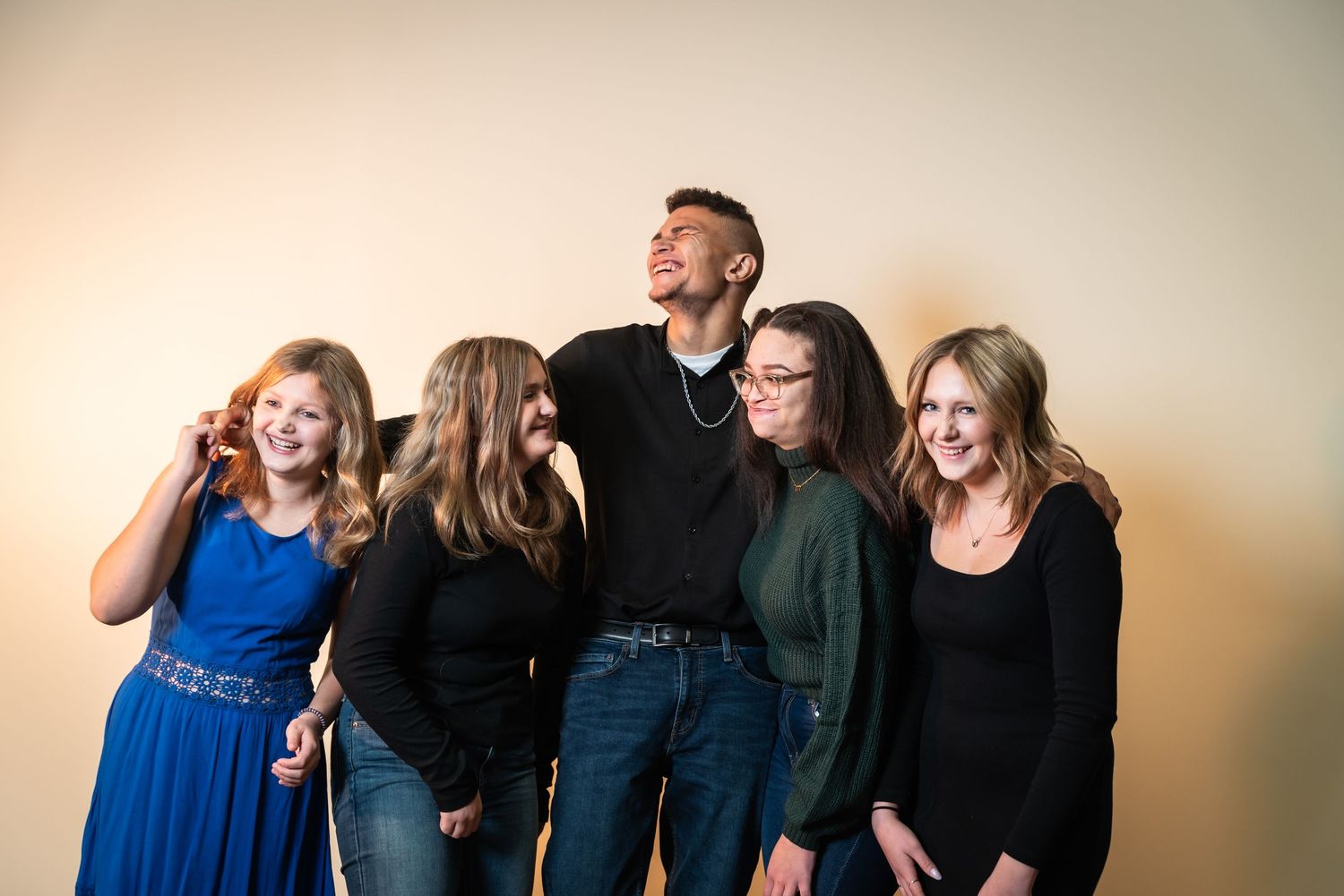 A group of friends laugh together while posing against a beige wall, wearing casual clothing in blue and black.