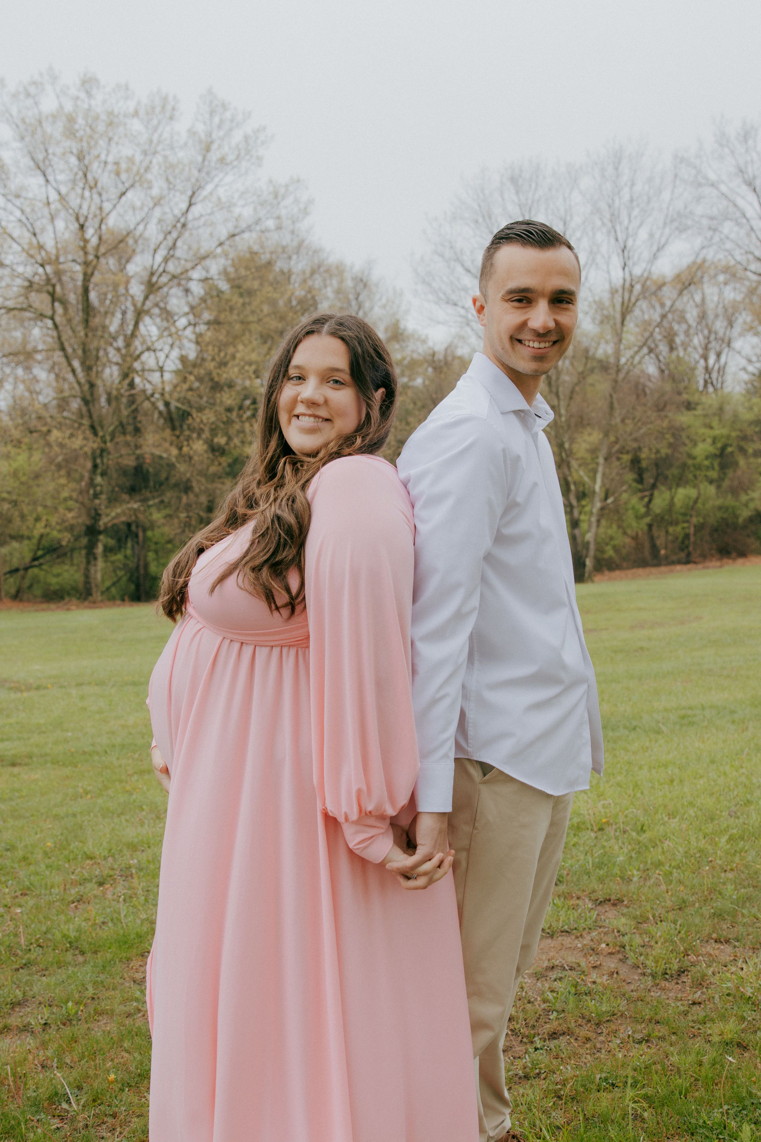 A couple poses back-to-back in a grassy field wearing pink and white outfits.