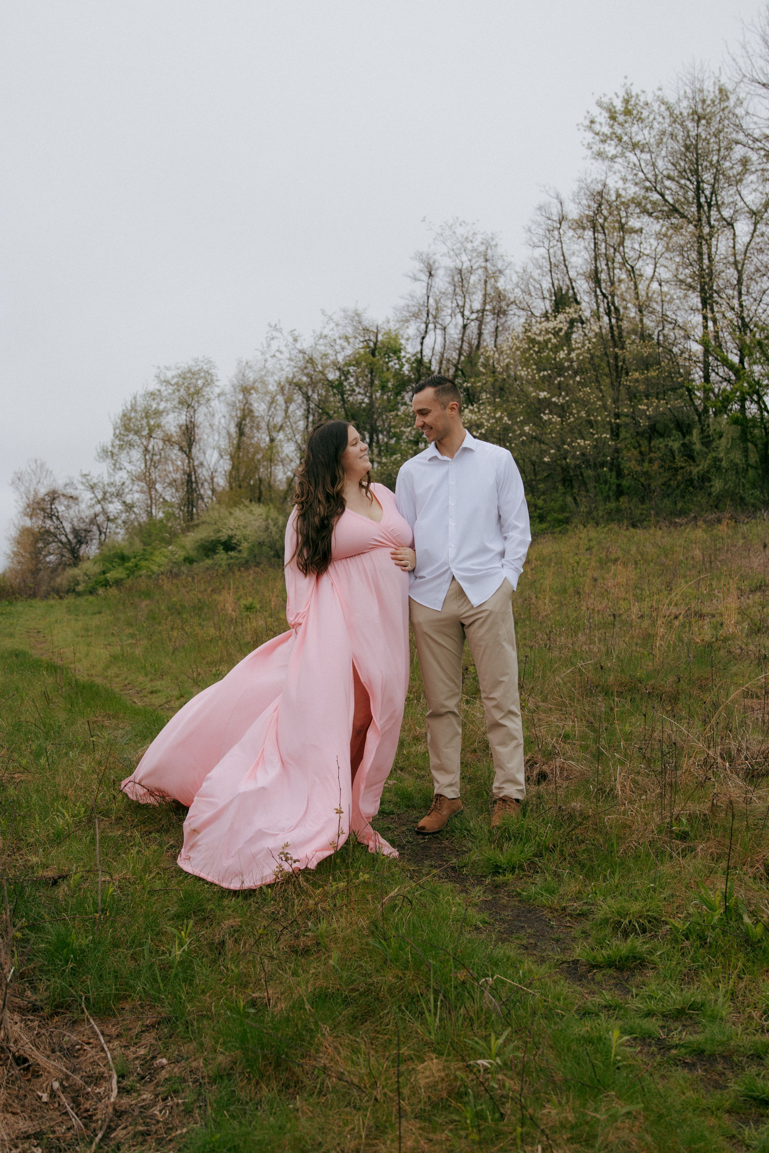 A couple poses romantically on a hillside, with a pink dress flowing in the breeze.