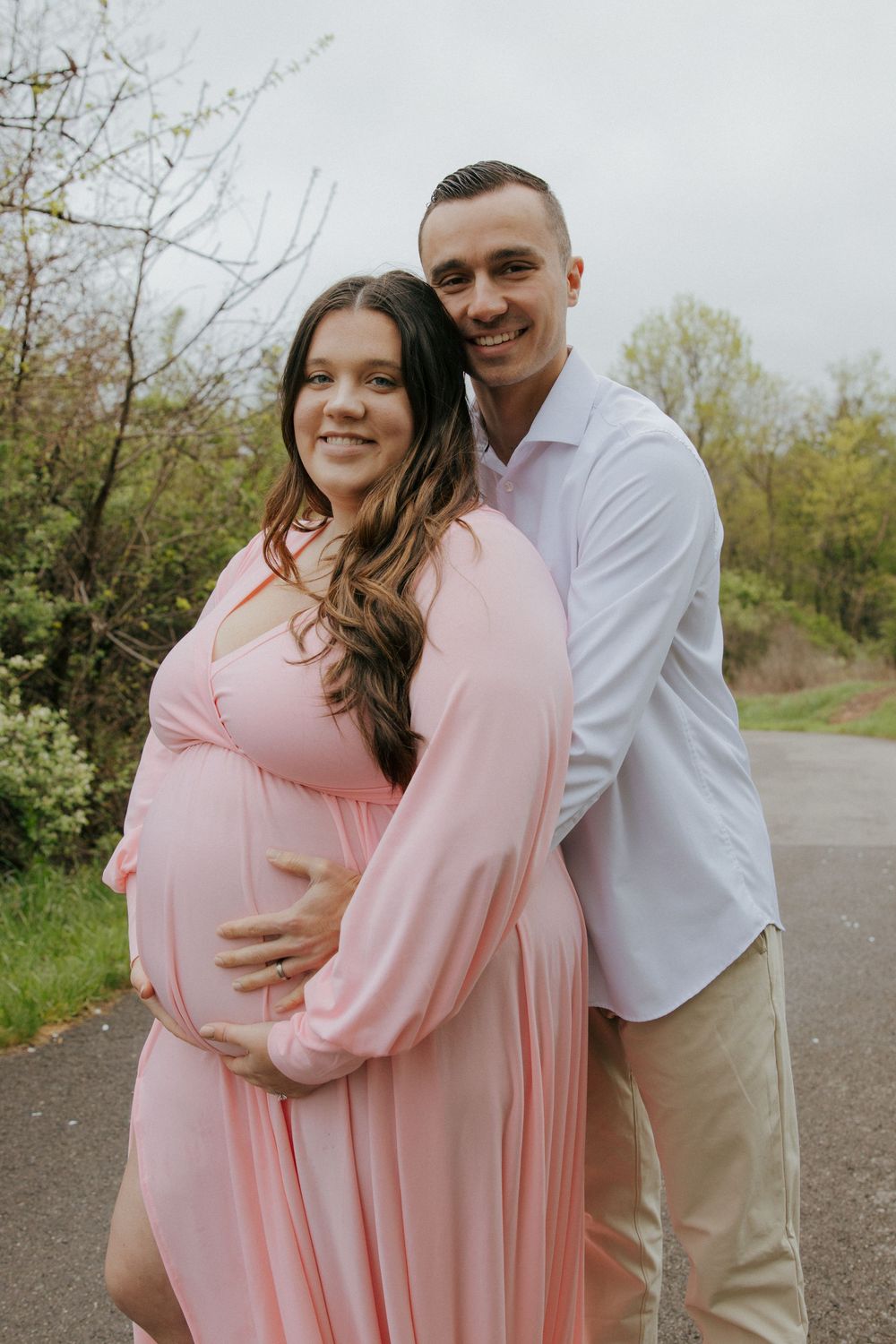 A couple embraces outdoors in spring, with the person in a flowing pink maternity dress showing a baby bump.