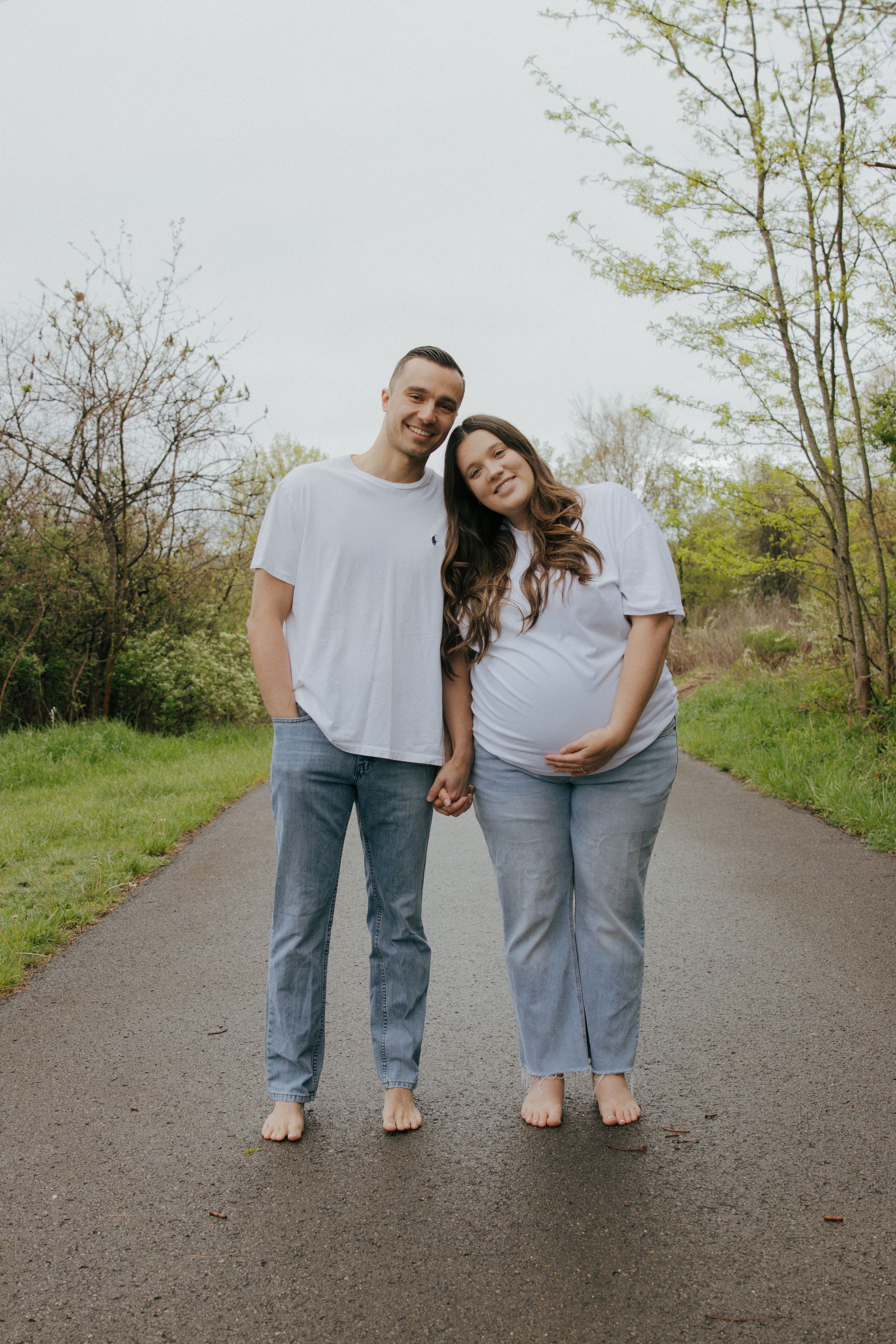 Couple wearing white shirts and jeans standing together on a trail surrounded by greenery.