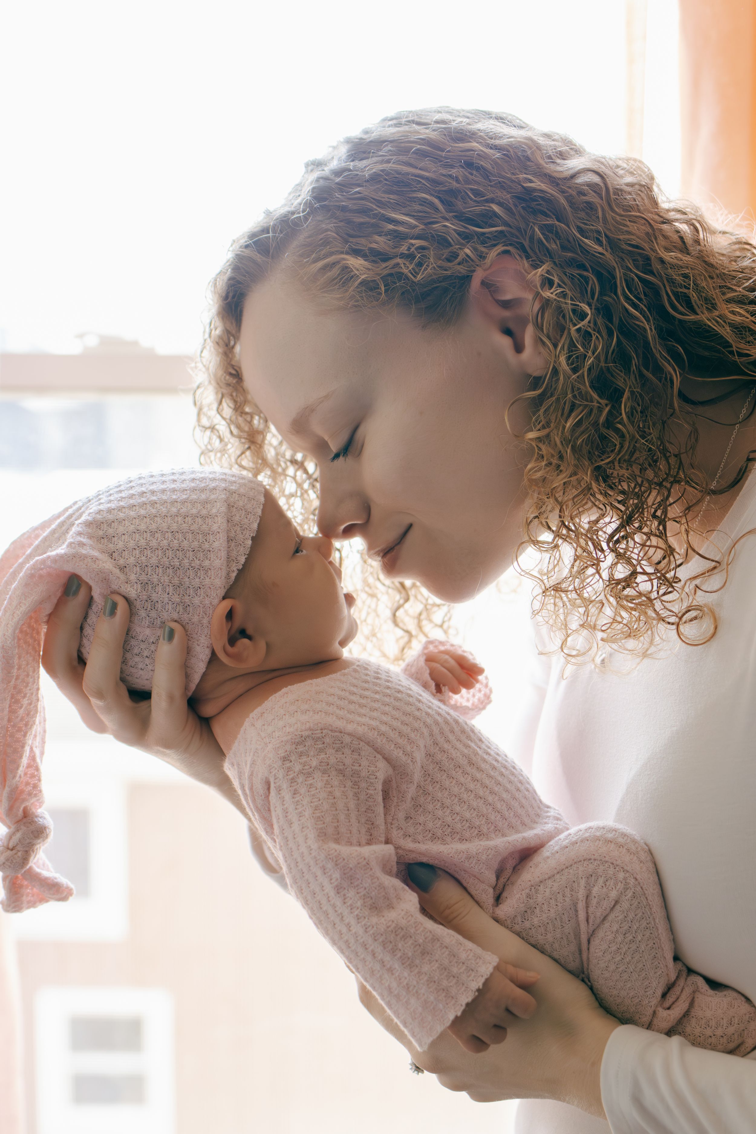 Close-up of loving parent nuzzling with baby in cozy pink sweater by window.