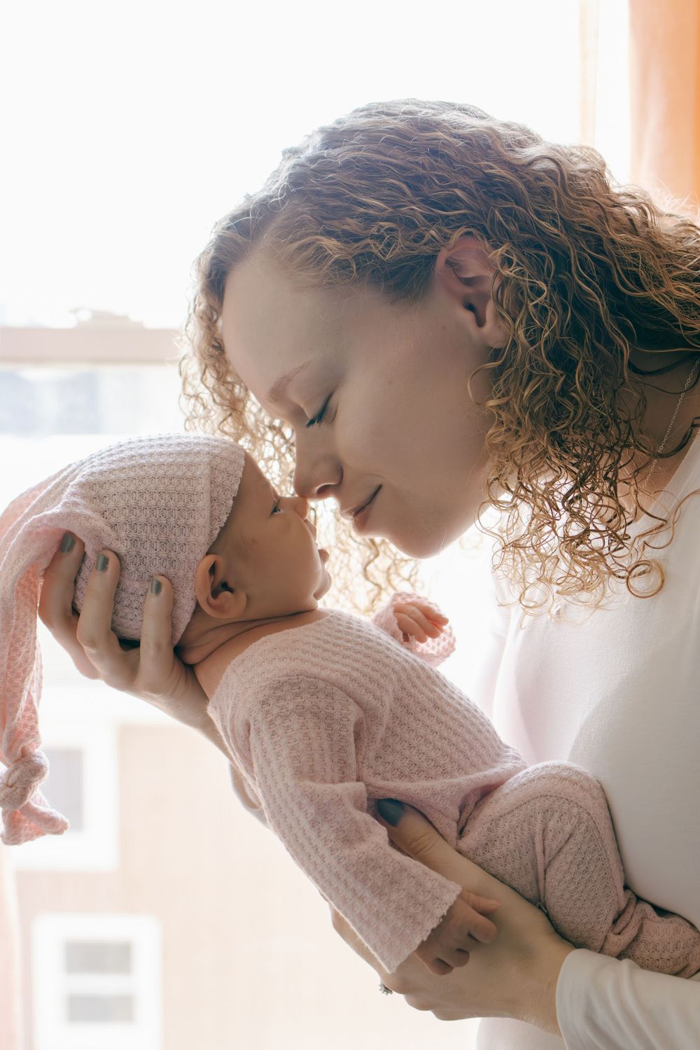 Close-up of loving parent nuzzling with baby in cozy pink sweater by window.