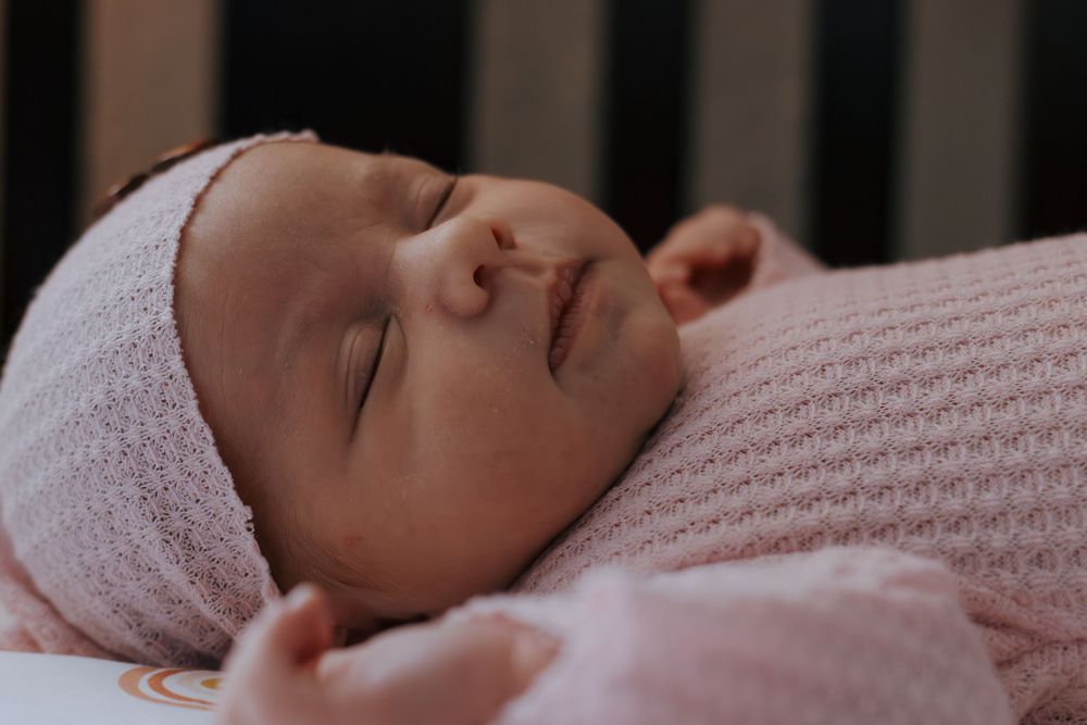 Baby peacefully napping in pink outfit viewed through dark crib bars.