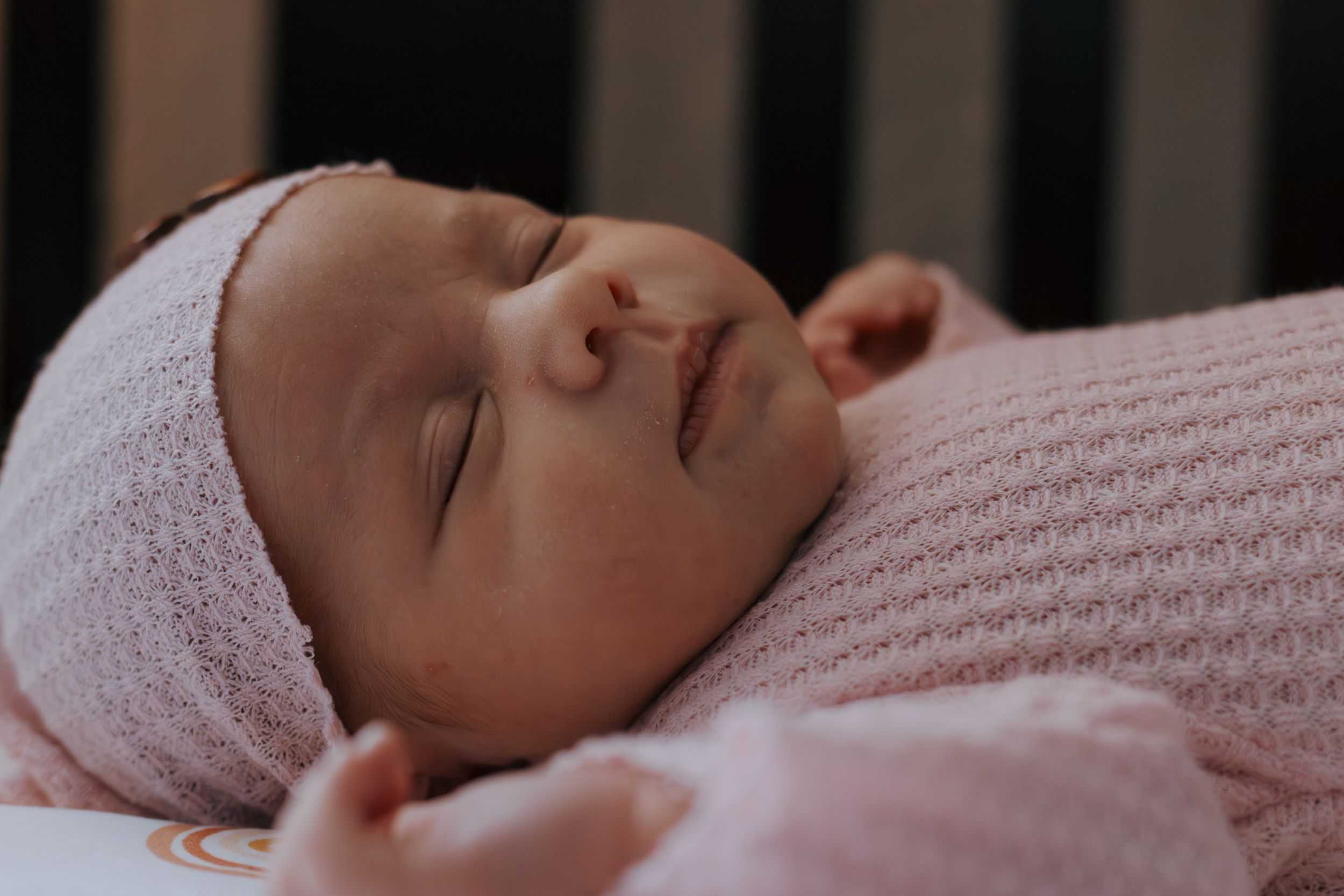 Baby peacefully napping in pink outfit viewed through dark crib bars.