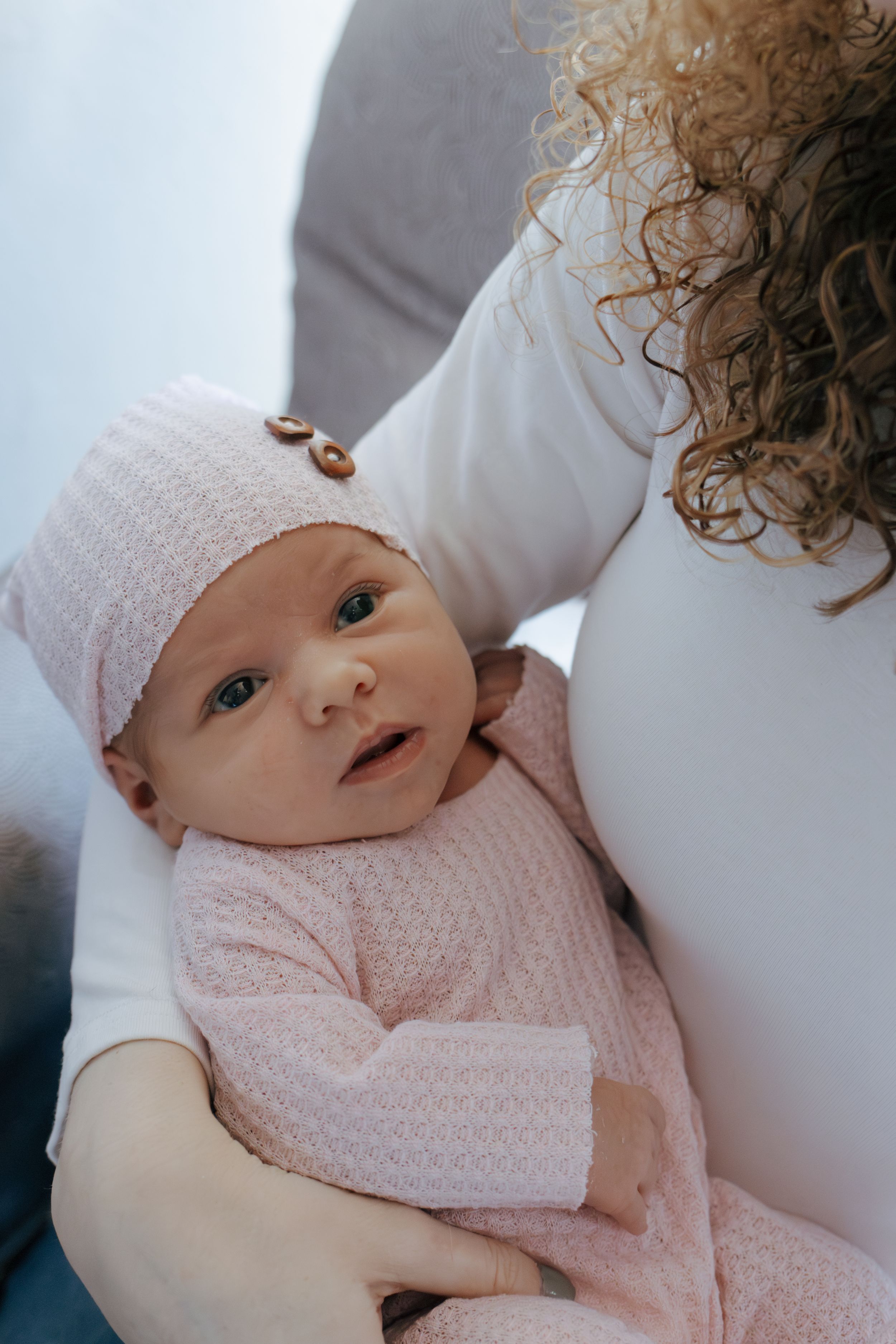 Sweet newborn baby wearing pink knit clothing and white hat rests peacefully on soft white bedding.