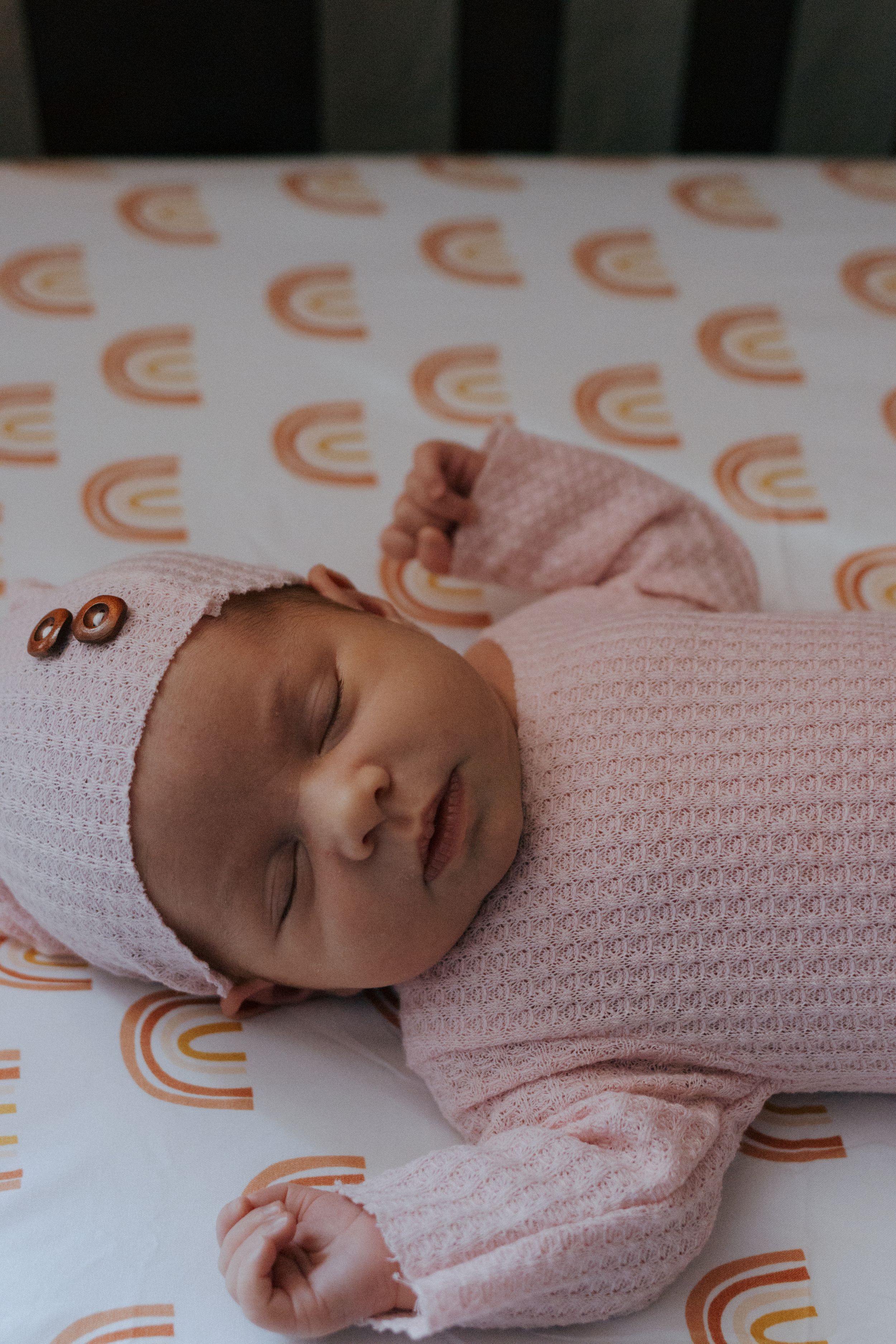 Peaceful baby resting on patterned sheet wearing light pink knitted outfit.
