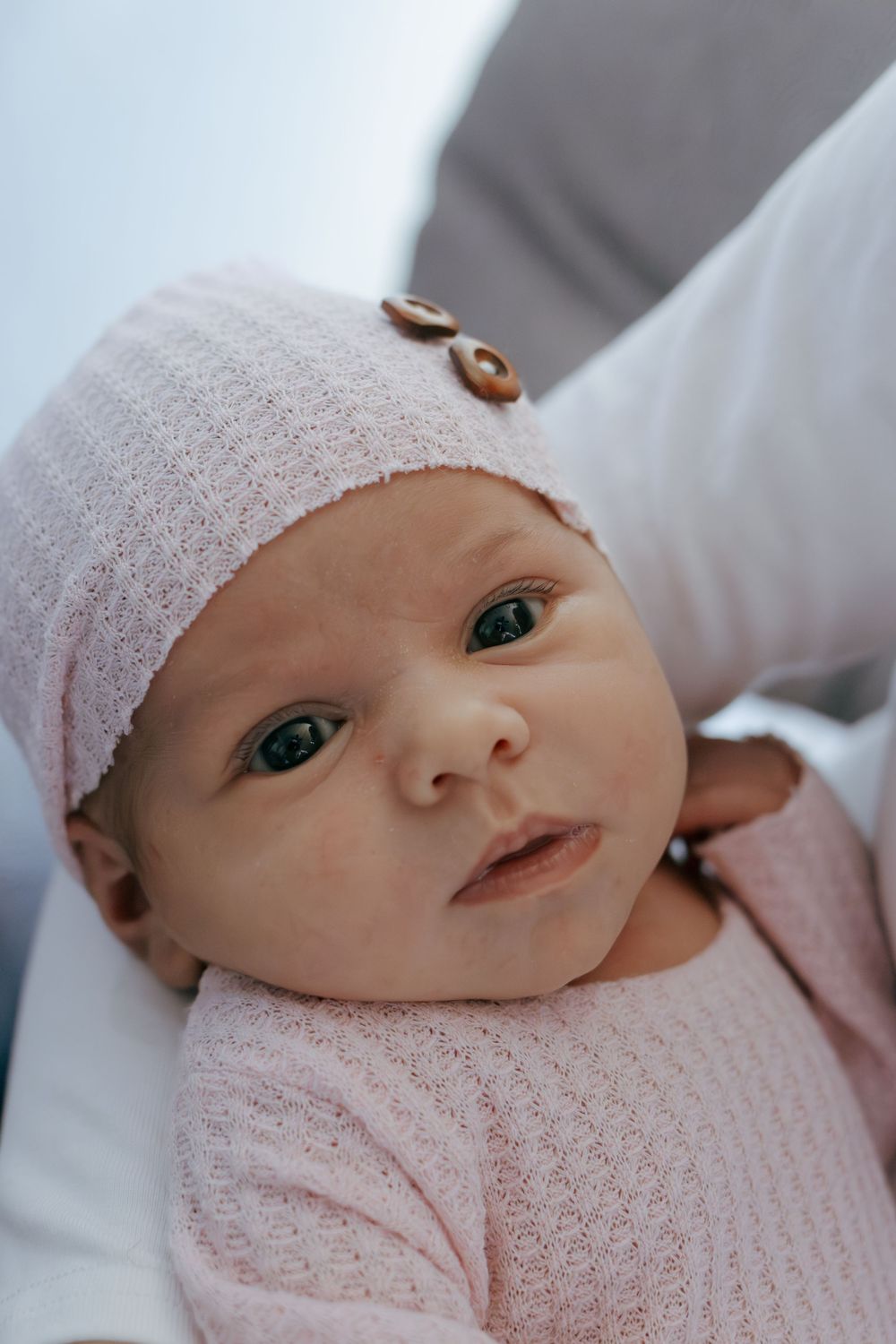 Close up of newborn baby in pink outfit and white knit hat with button detail.