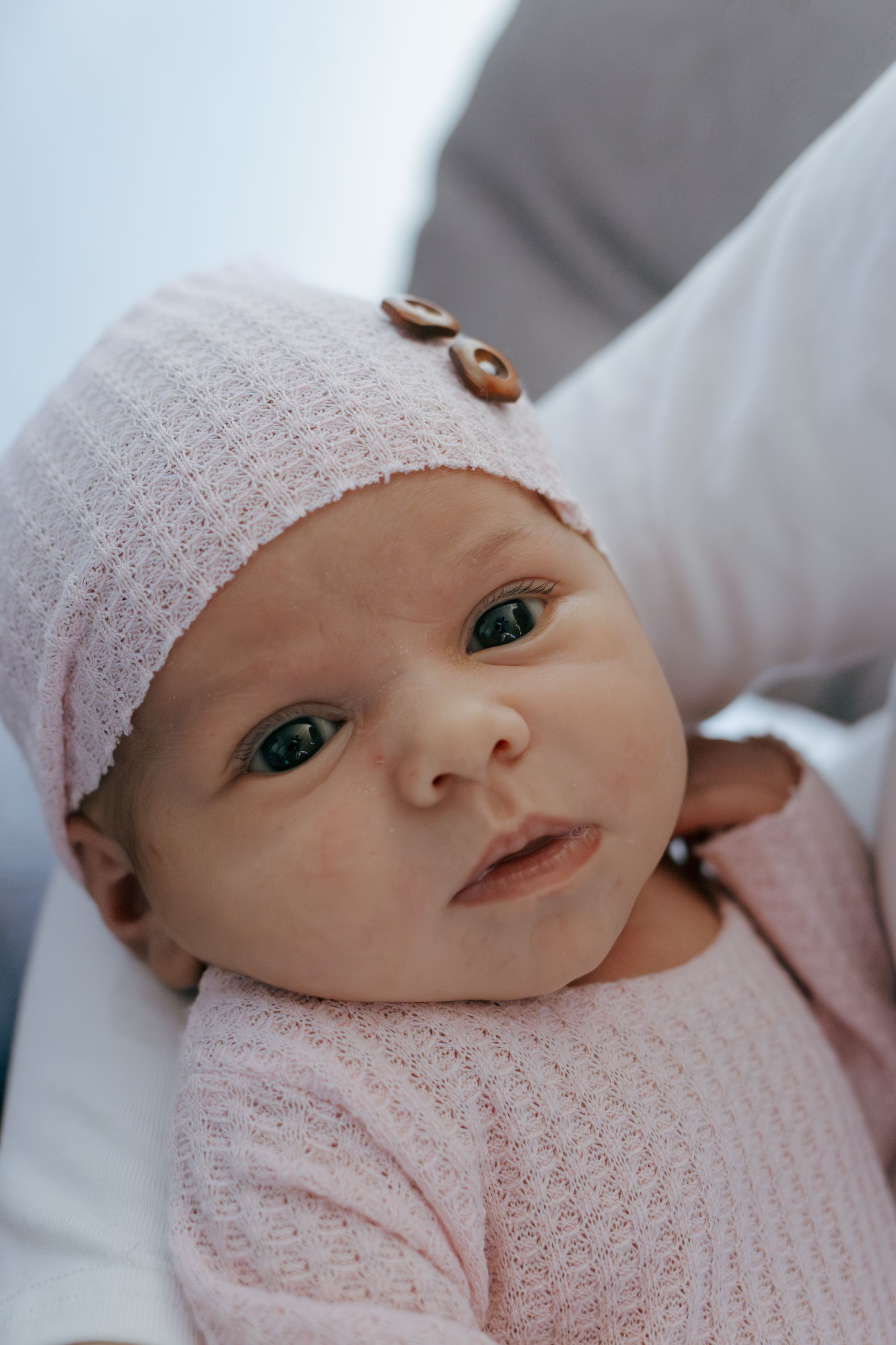 Close up of newborn baby in pink outfit and white knit hat with button detail.
