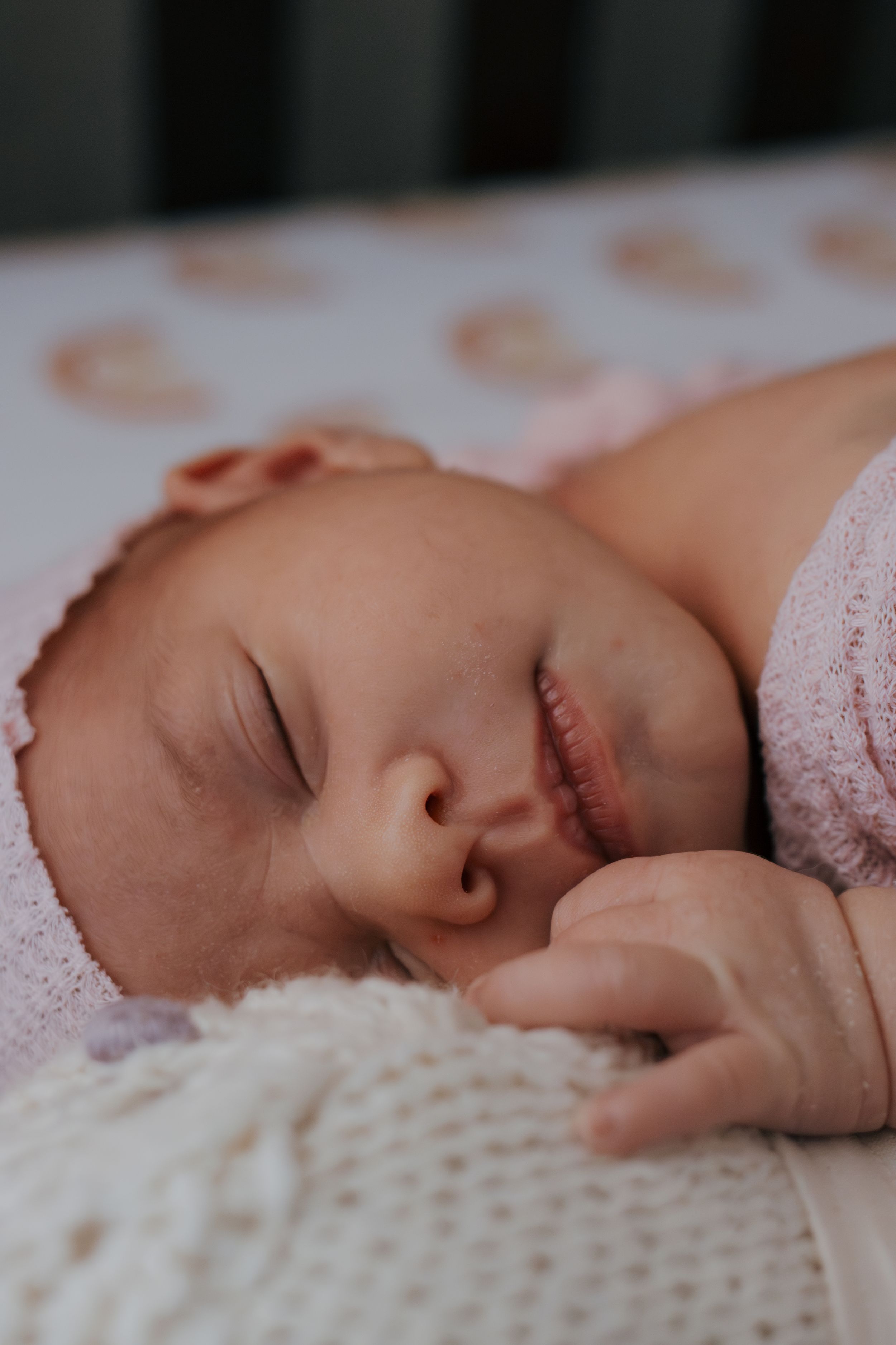 Sleeping newborn baby rests peacefully on white textured blanket wearing delicate pink clothing.
