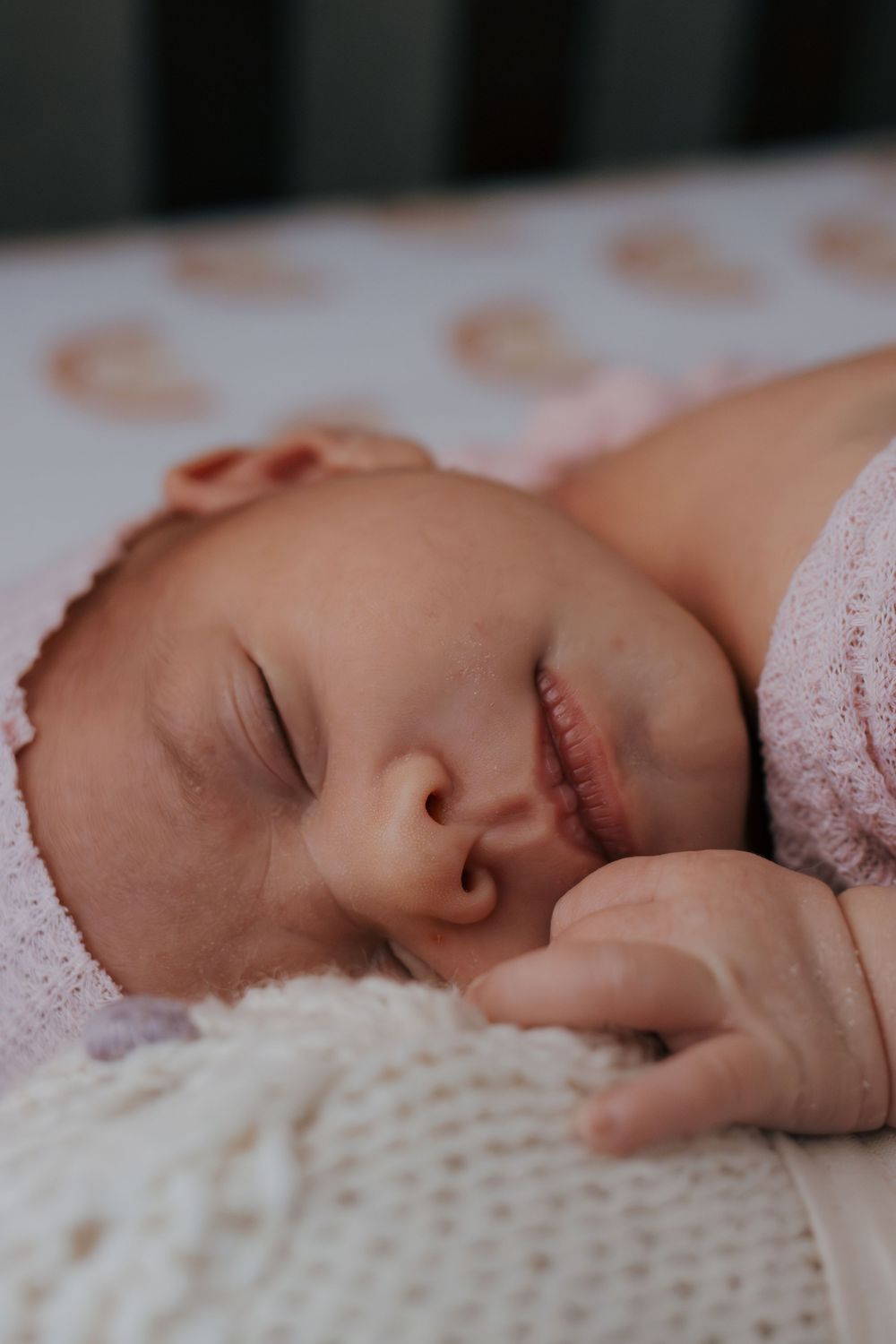 Sleeping newborn baby rests peacefully on white textured blanket wearing delicate pink clothing.