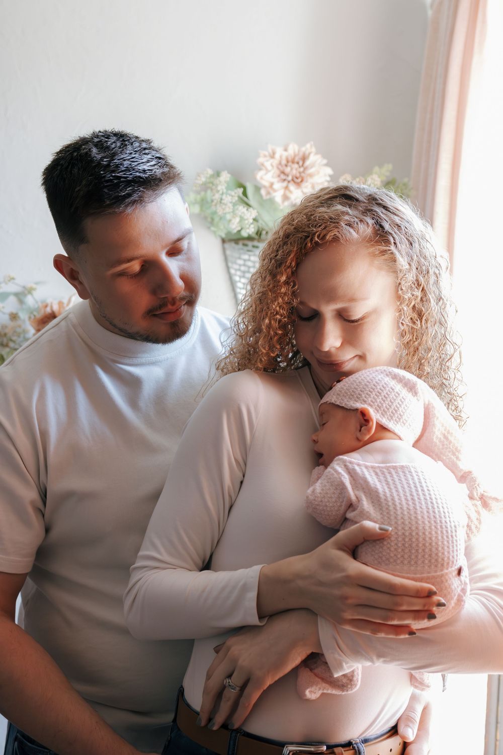 Tender family moment as parents embrace their newborn in soft natural window light.