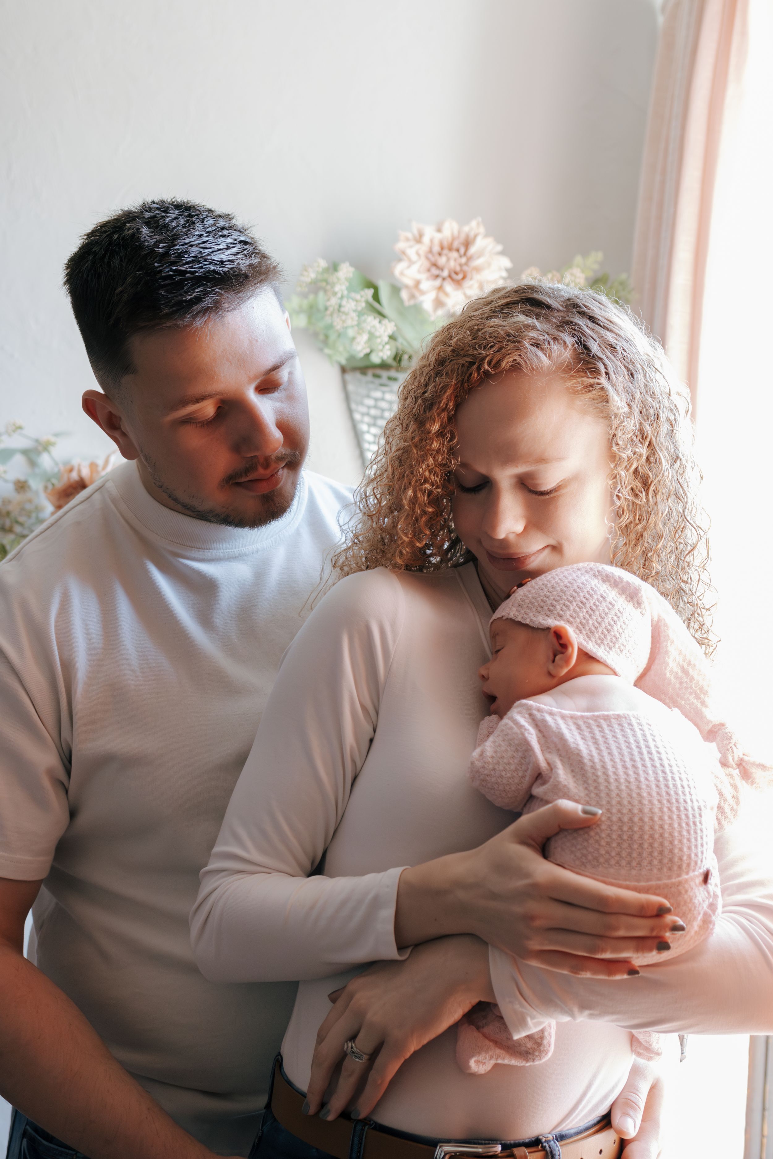 Tender family moment as parents embrace their newborn in soft natural window light.