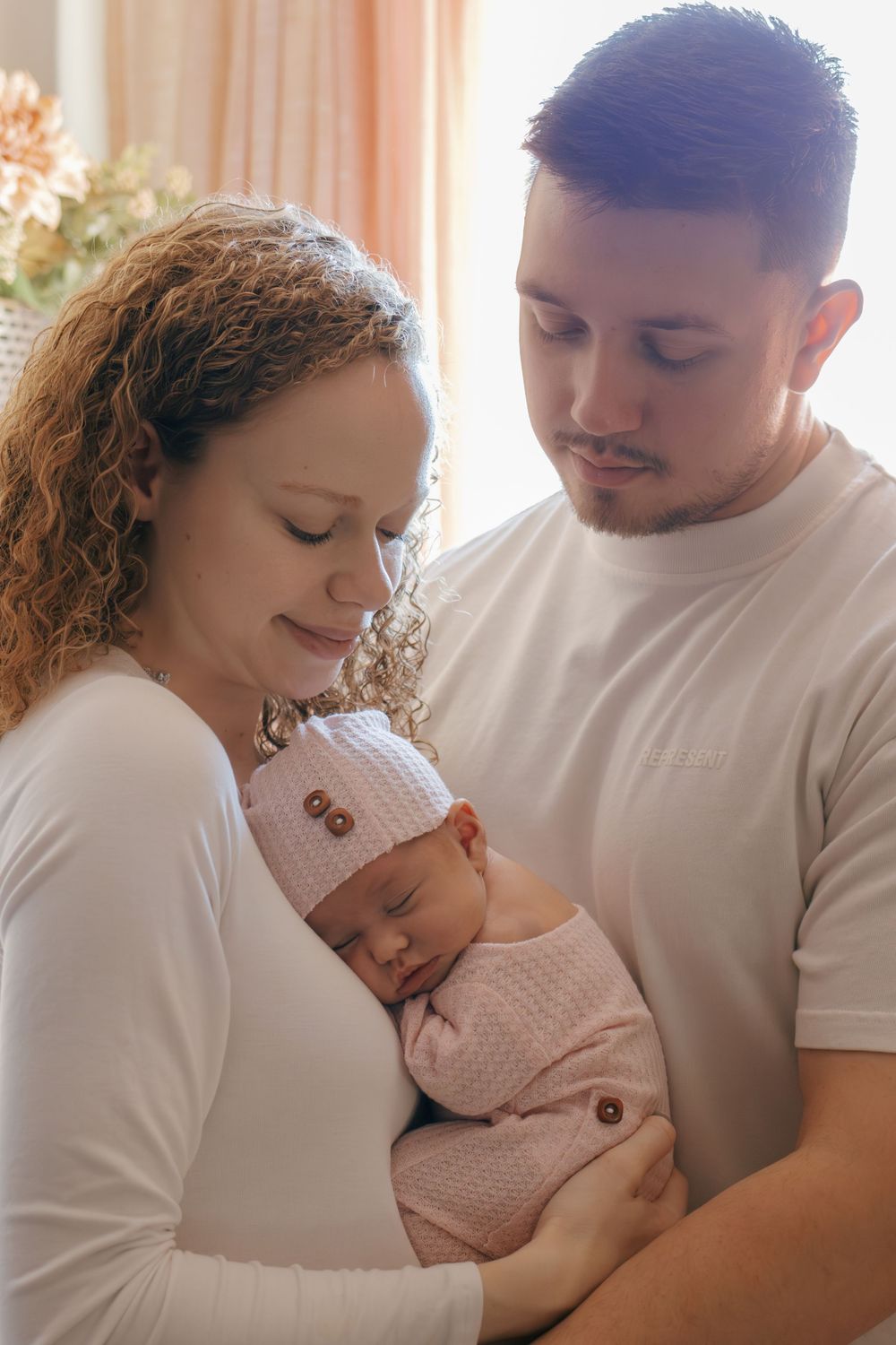 Intimate family portrait with parents holding their sleeping newborn in pink against window light.