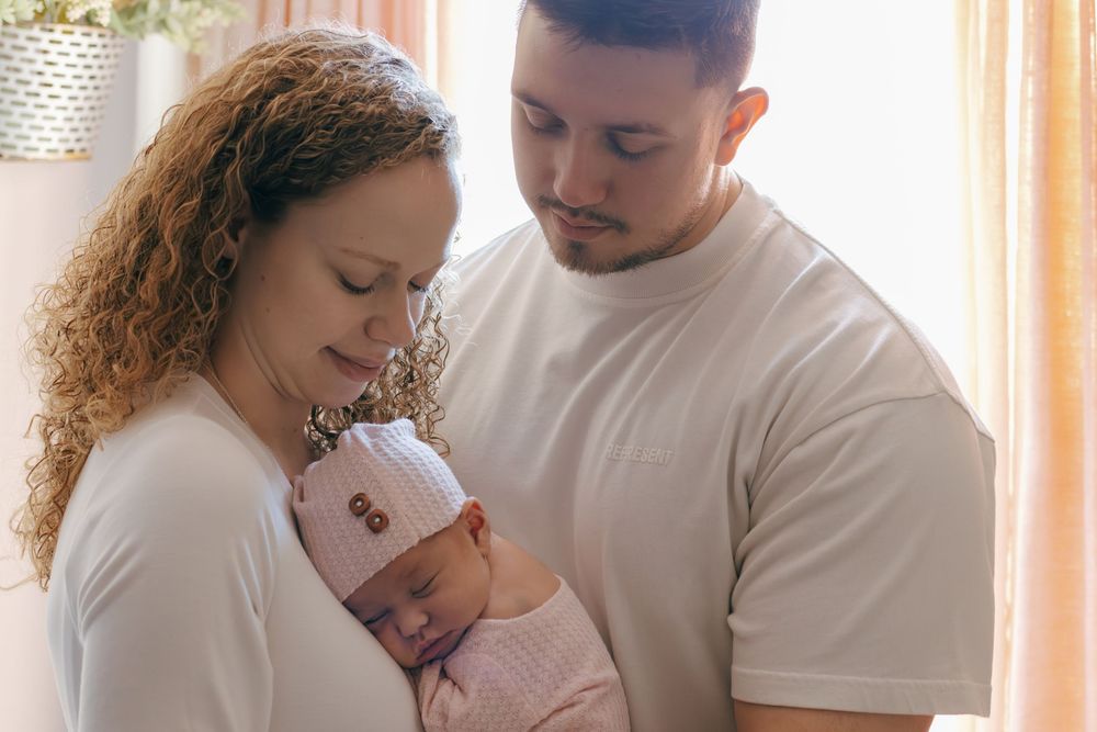 Gentle family moment captured in soft window lighting with parents cradling their newborn.