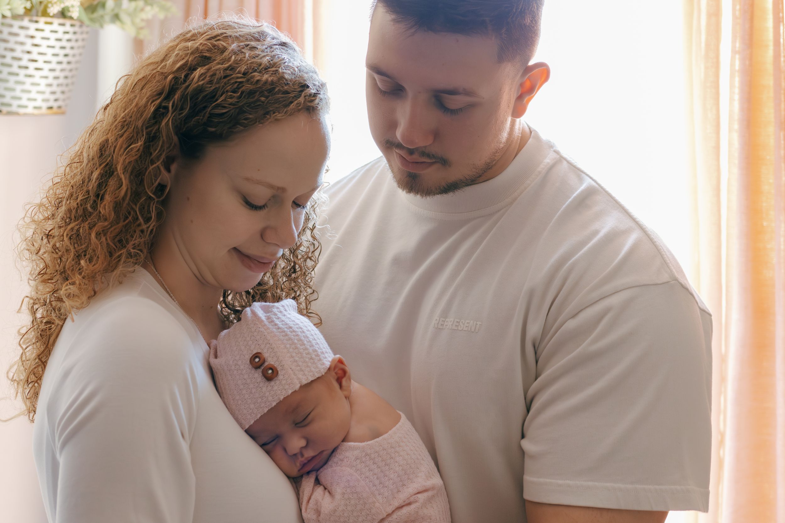 Gentle family moment captured in soft window lighting with parents cradling their newborn.