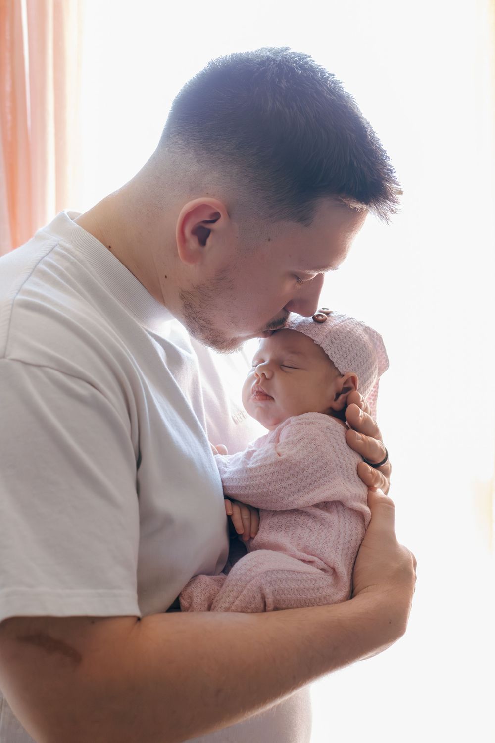 Father in white shirt tenderly kisses newborn baby wearing pink in bright window light.