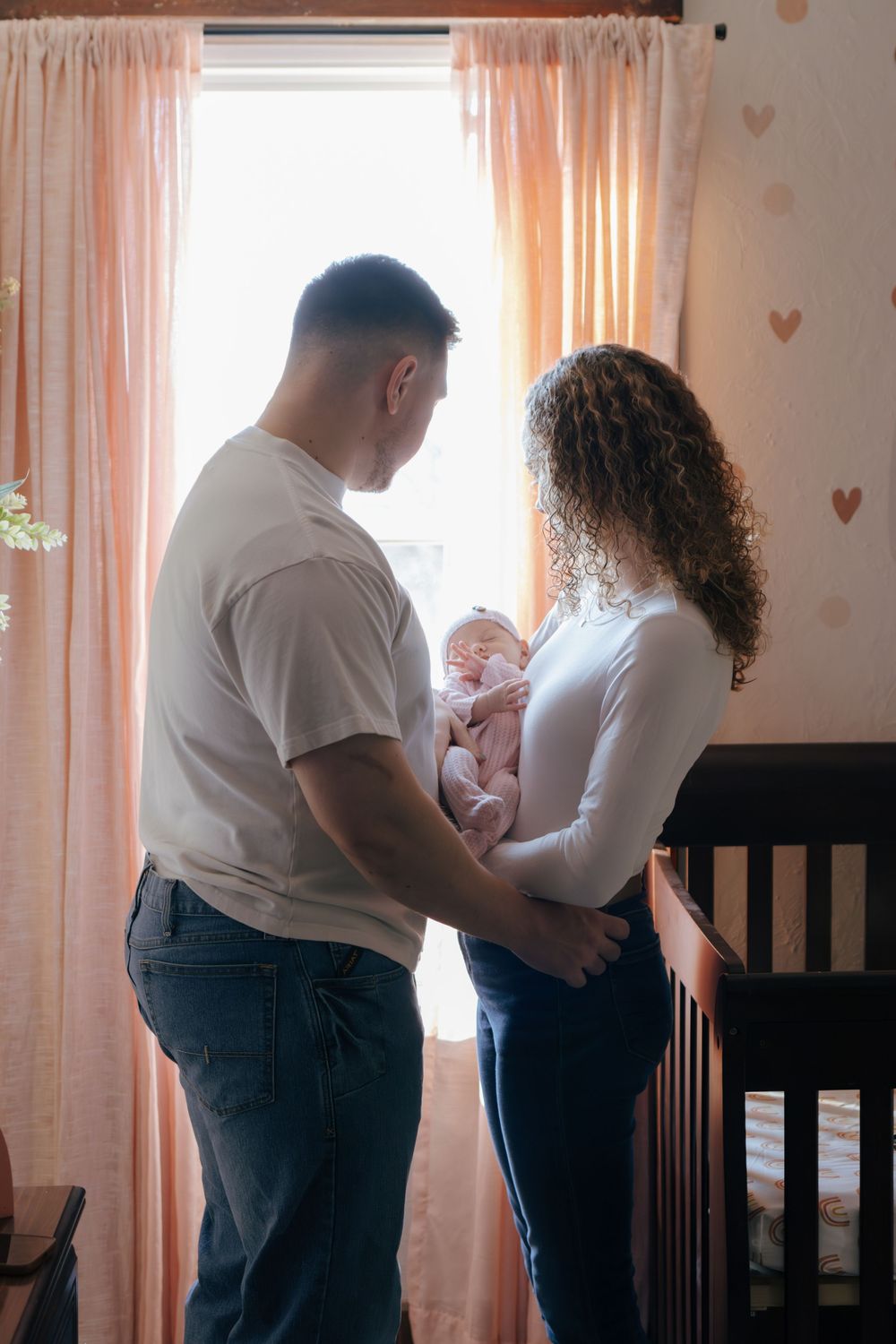 Parents in white and denim stand by nursery windows with their newborn.