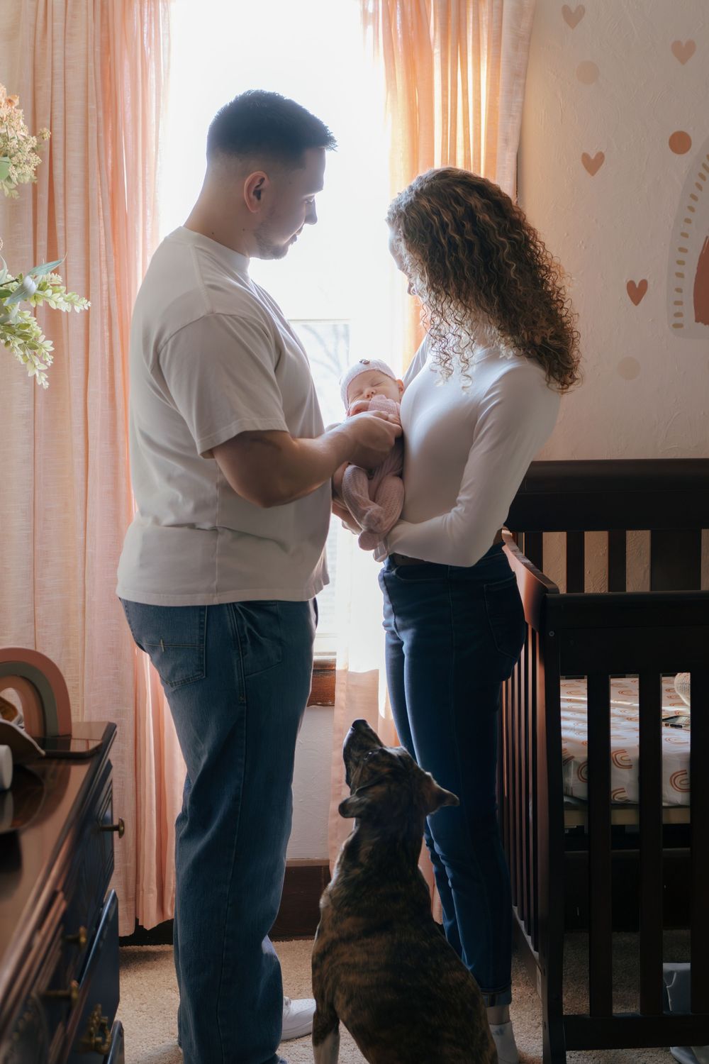 Couple stands near a dark wooden crib holding their newborn in a room with peach curtains.