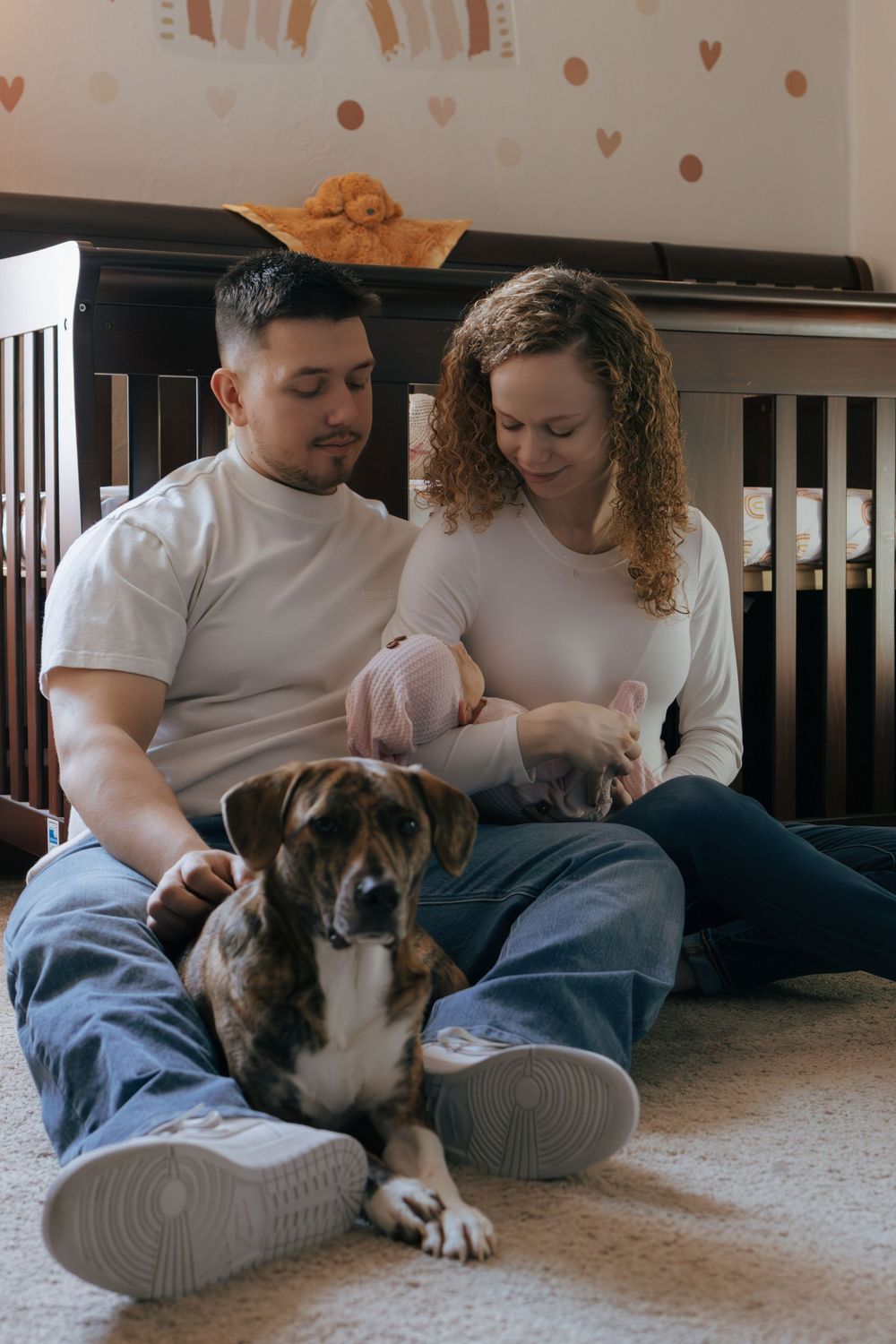 Family moment with dog as couple relaxes on carpeted floor by dark furniture.