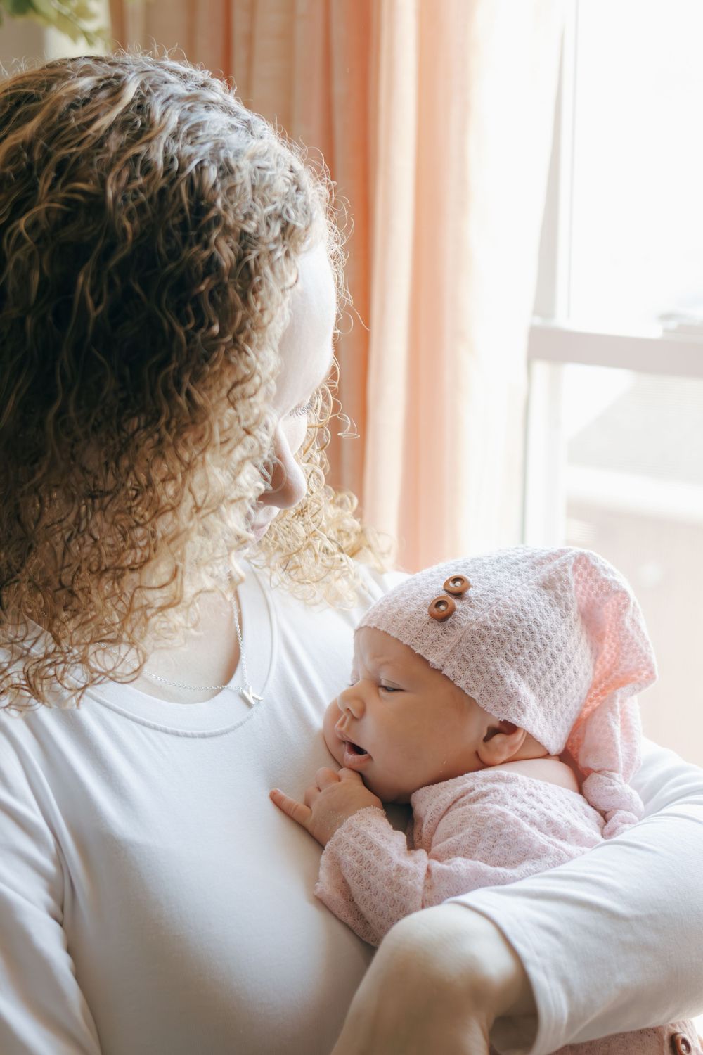 Intimate portrait of baby nestled against caregiver in sunlit room with peach curtains.