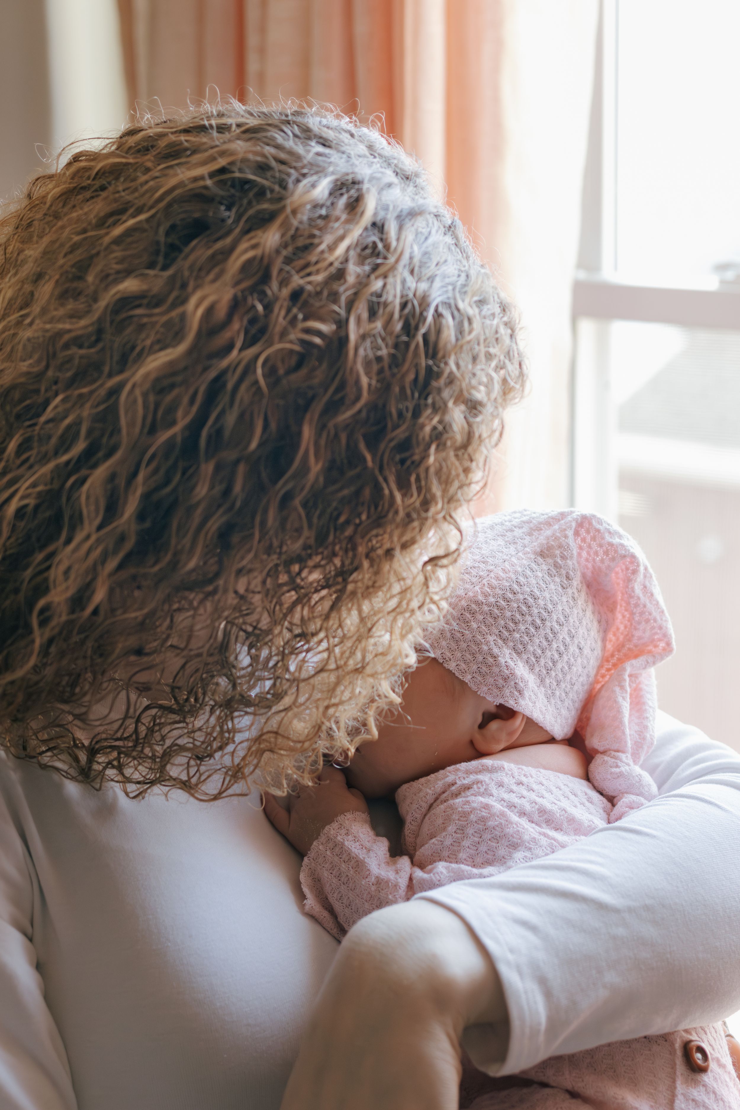 Gentle embrace between caregiver with curly hair and infant in pink knit clothing.