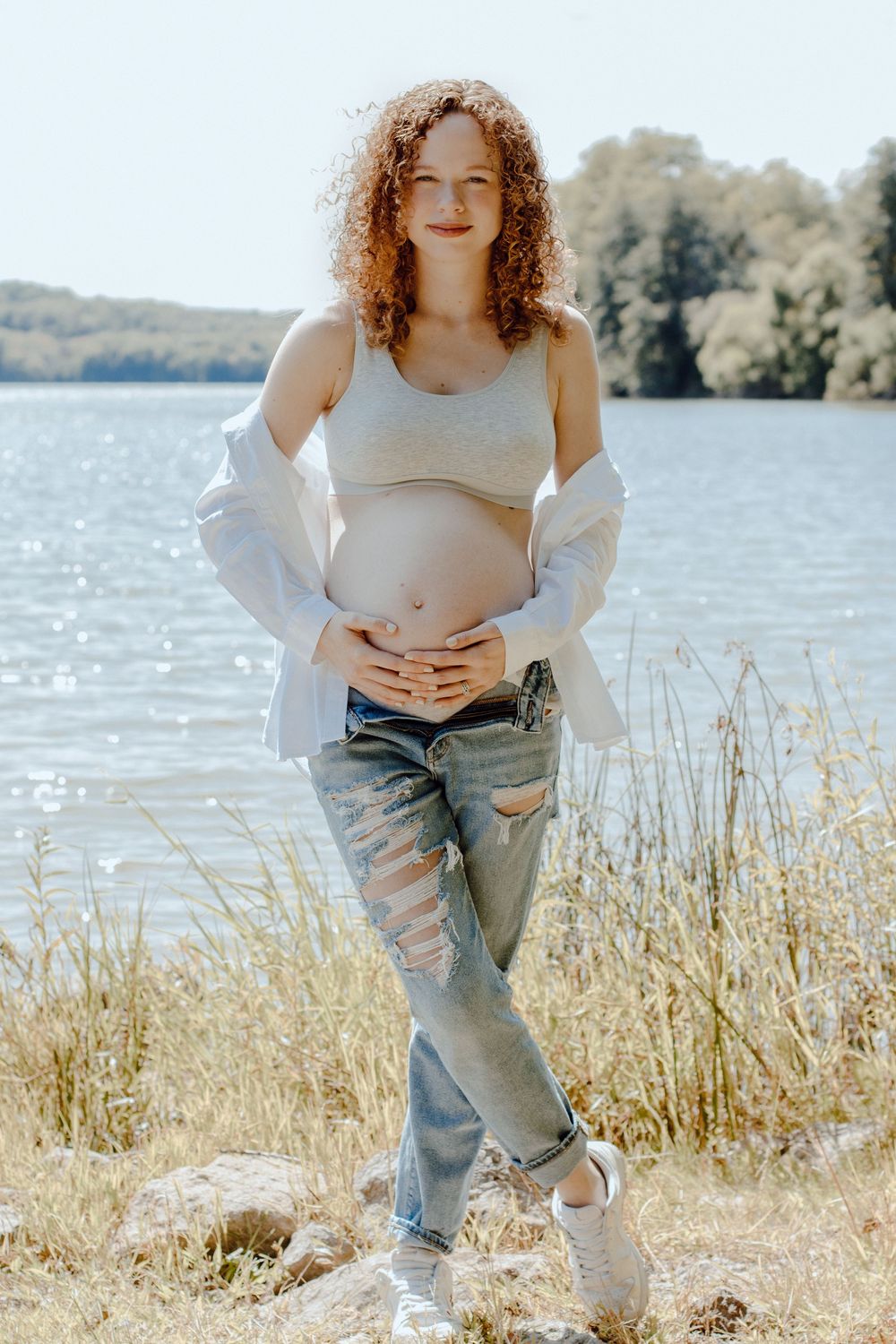 Person in white top and distressed jeans poses by lake during maternity photoshoot.