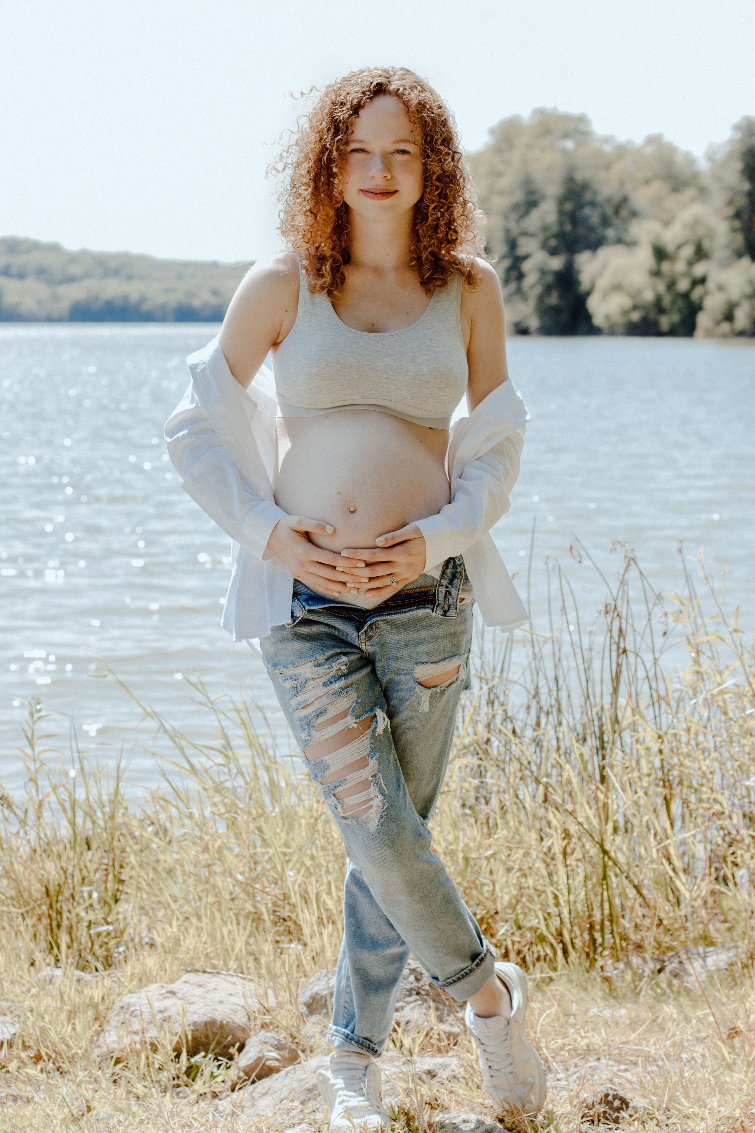 Person in white top and distressed jeans poses by lake during maternity photoshoot.
