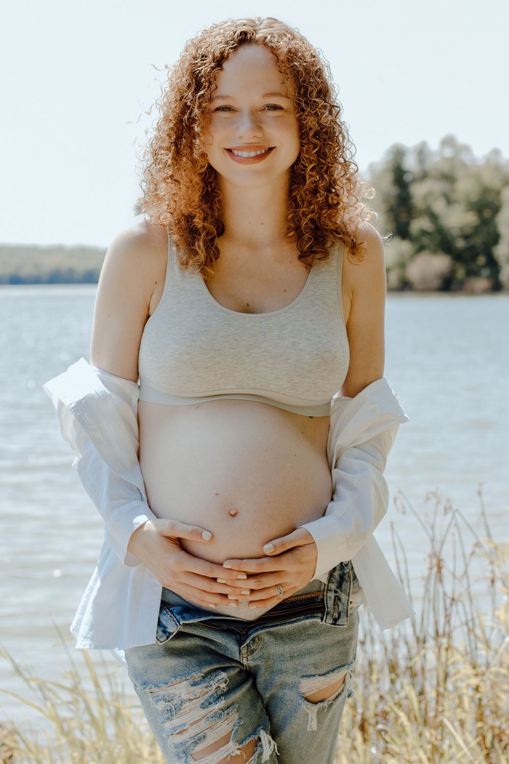 Maternity portrait by lake showing pregnant belly with sunlit water in background.