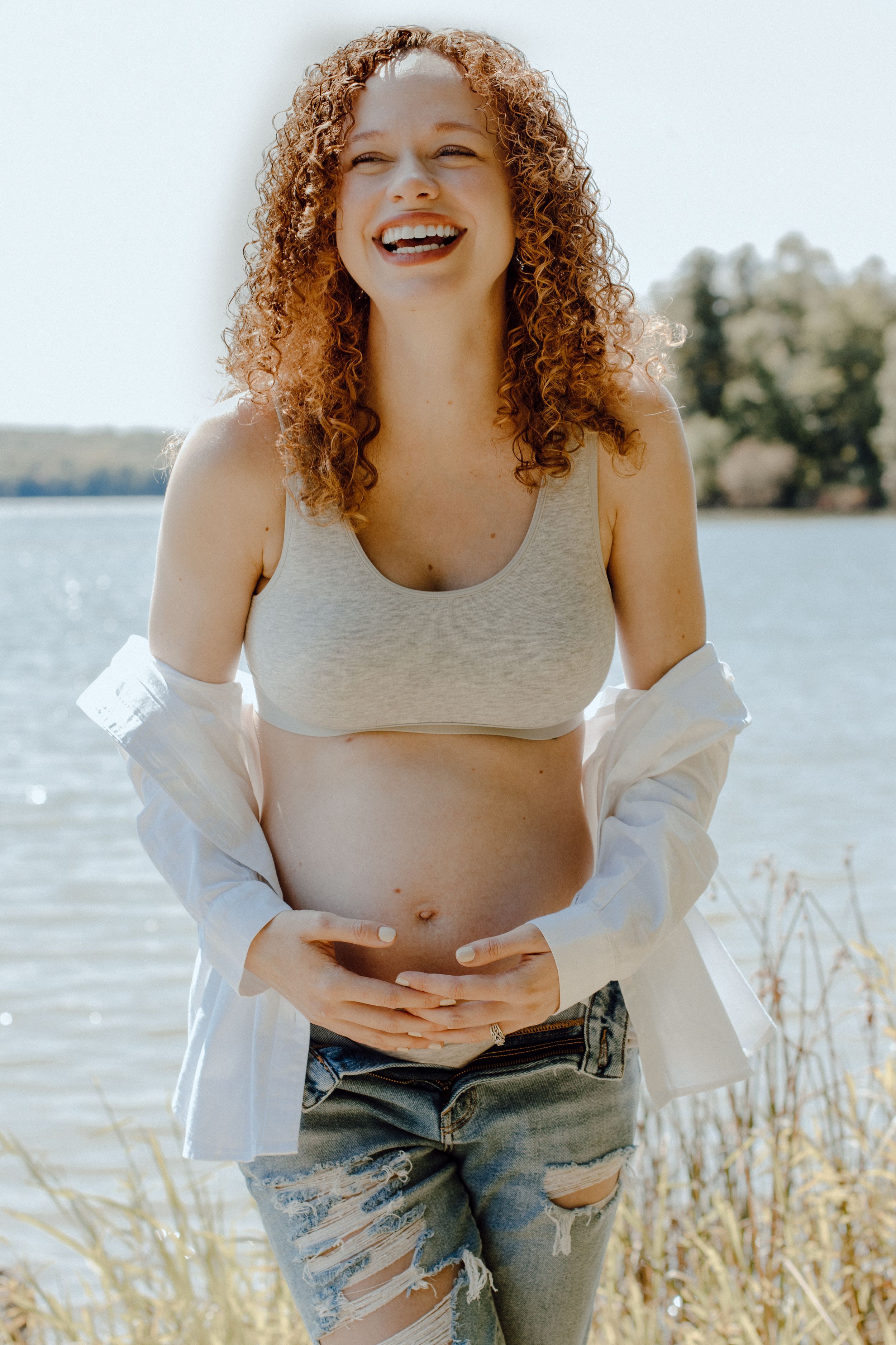 Candid maternity photoshoot by a lake showing a joyful moment in distressed jeans and grey crop top.
