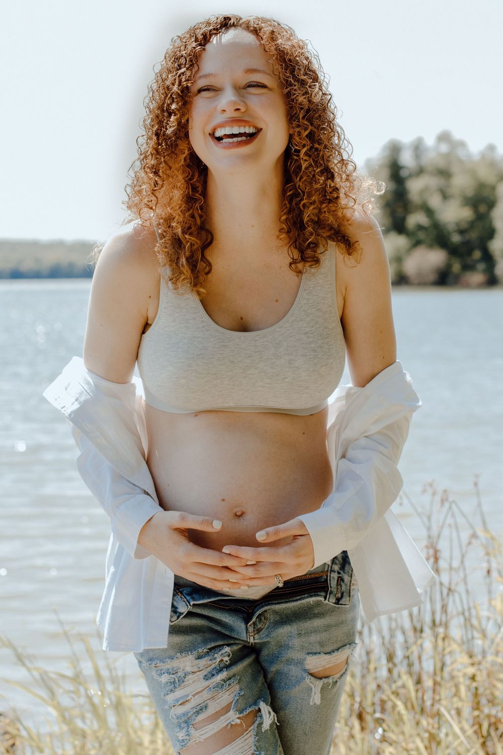 Candid maternity photoshoot by a lake showing a joyful moment in distressed jeans and grey crop top.