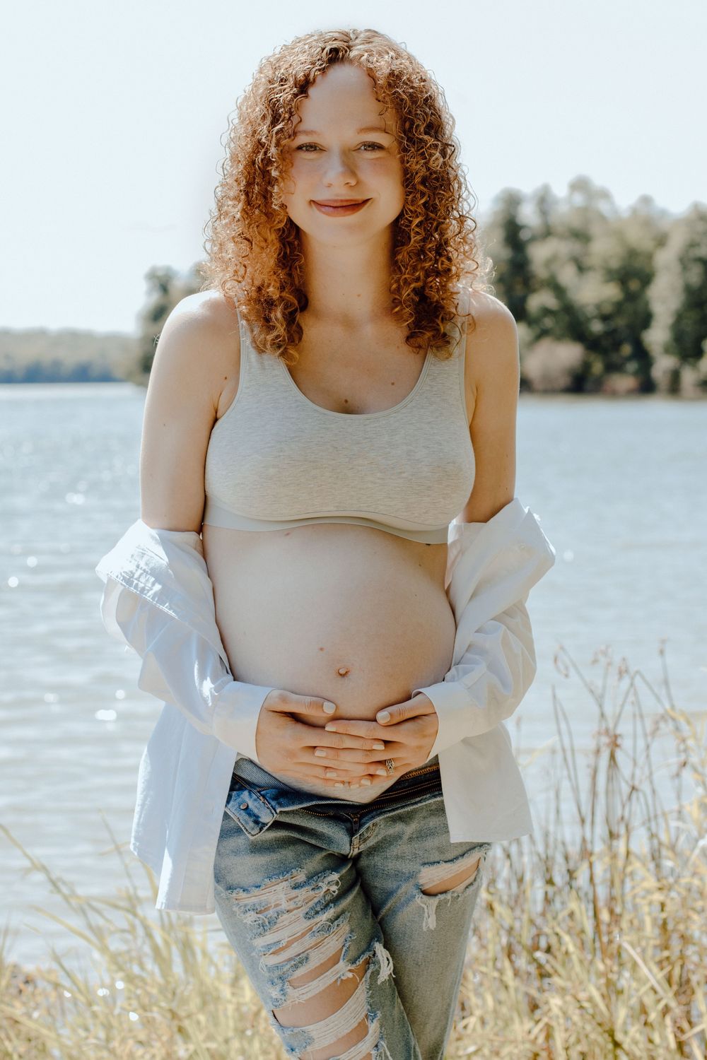 Casual maternity portraits taken near water featuring ripped jeans and a white button-down shirt.