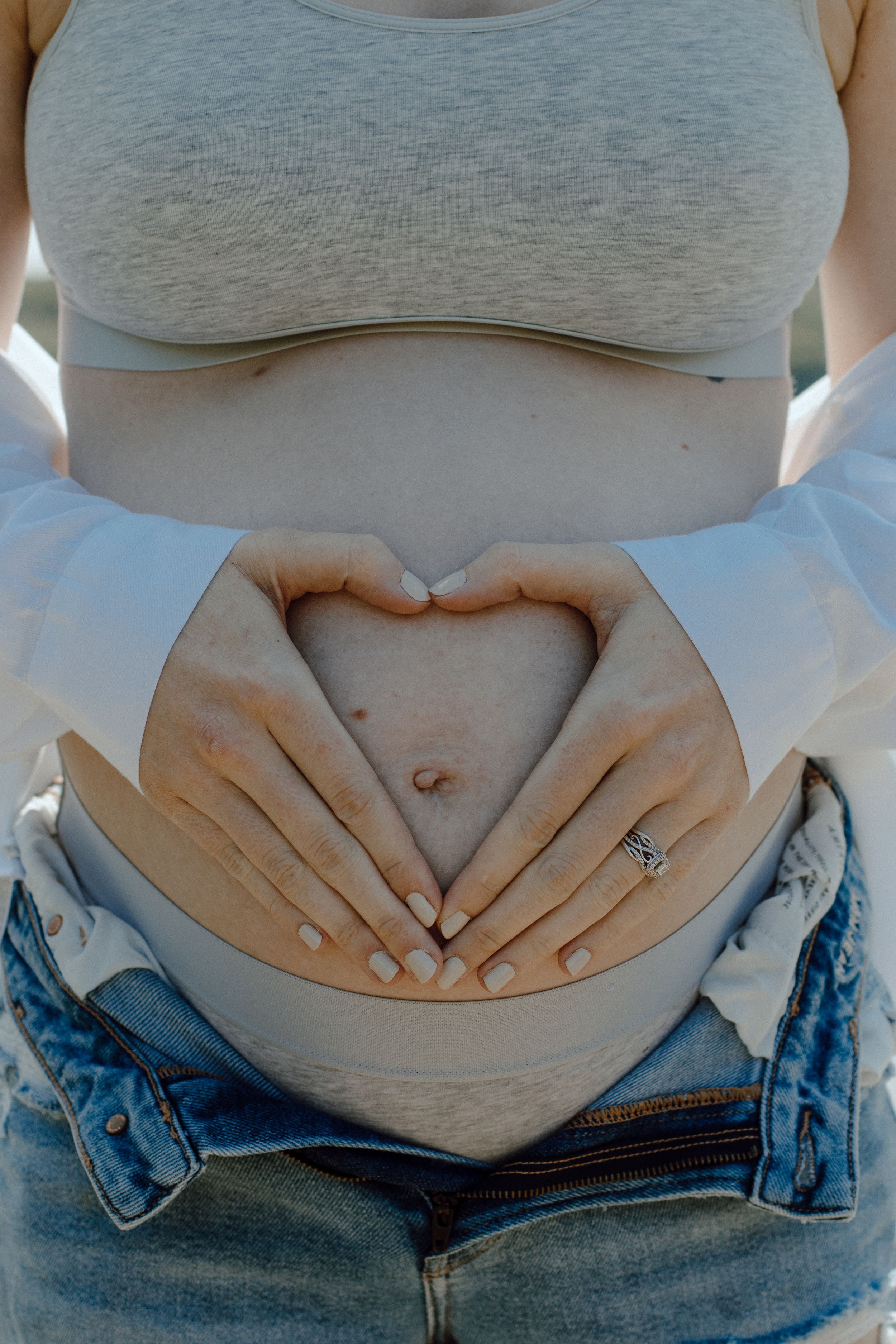 Close-up of hands forming heart shape over pregnant belly against grey top and distressed denim.