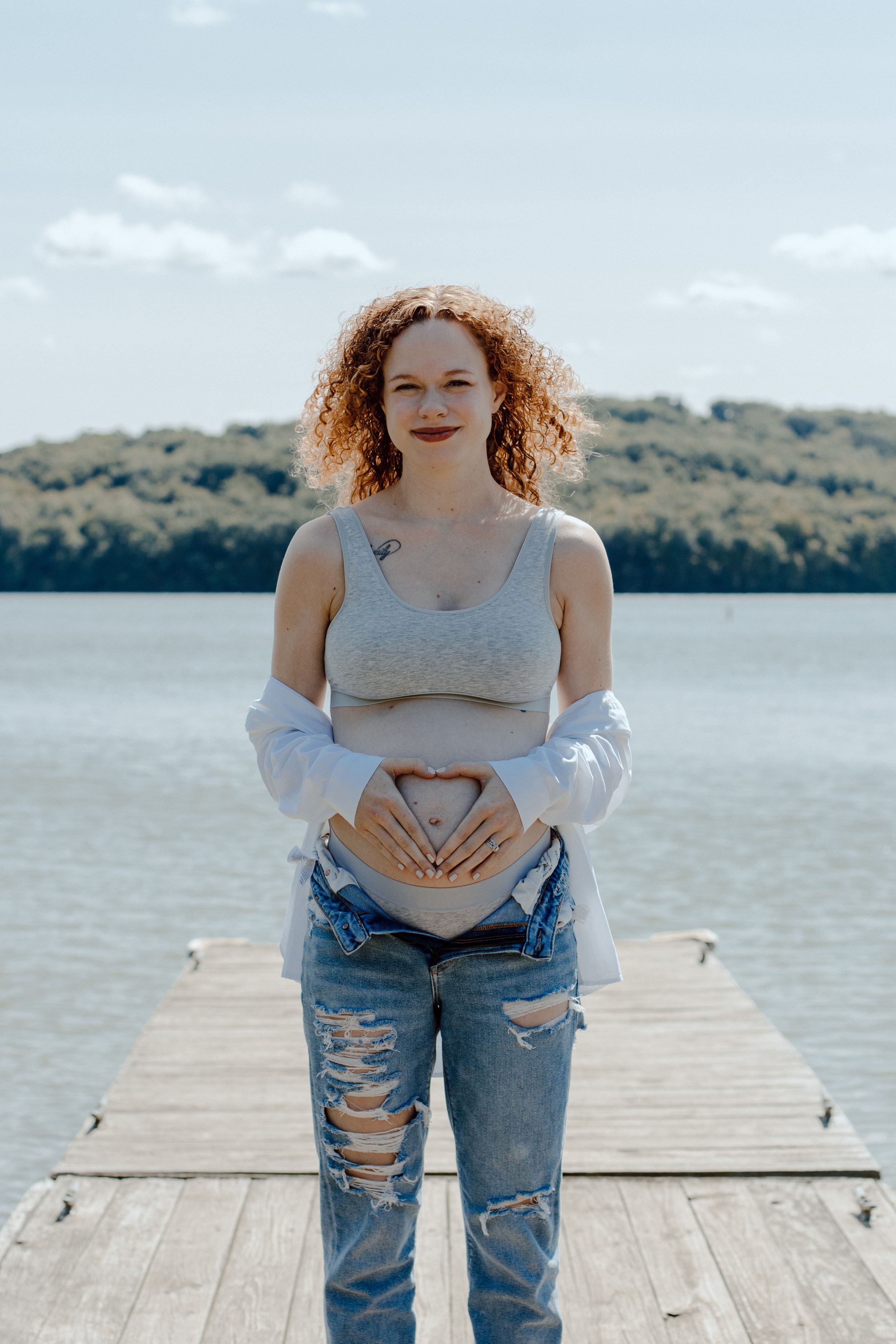 Maternity photos on wooden dock with lake and forest backdrop showing playful poses.