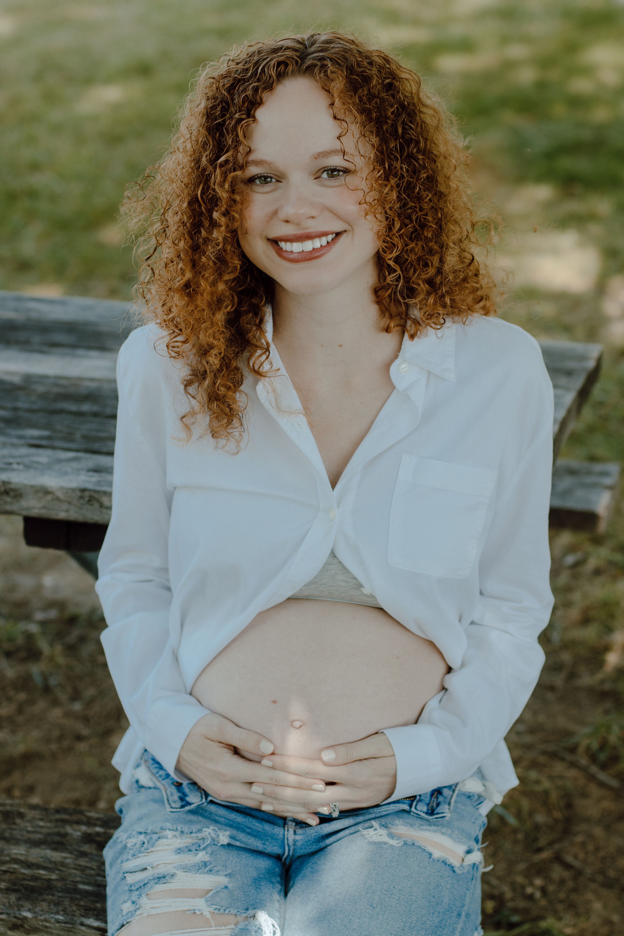 Person in white button-down shirt and jeans sitting outdoors with curly hair and bright smile.
