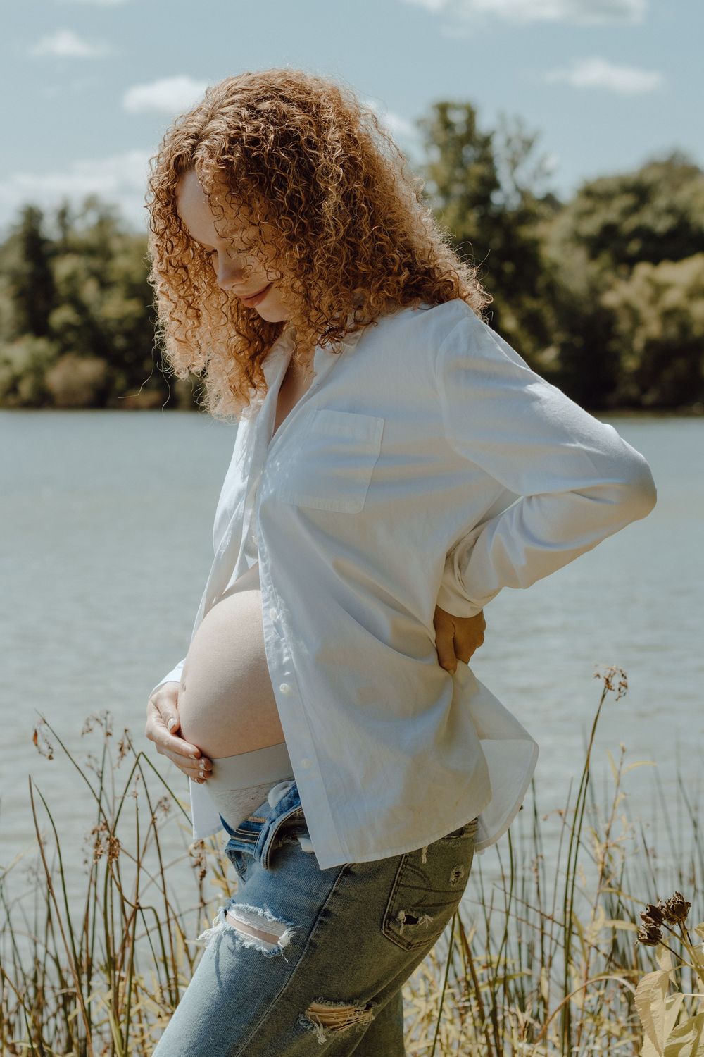 Person with curly hair in white sweater by lakeside with tall grass in foreground.