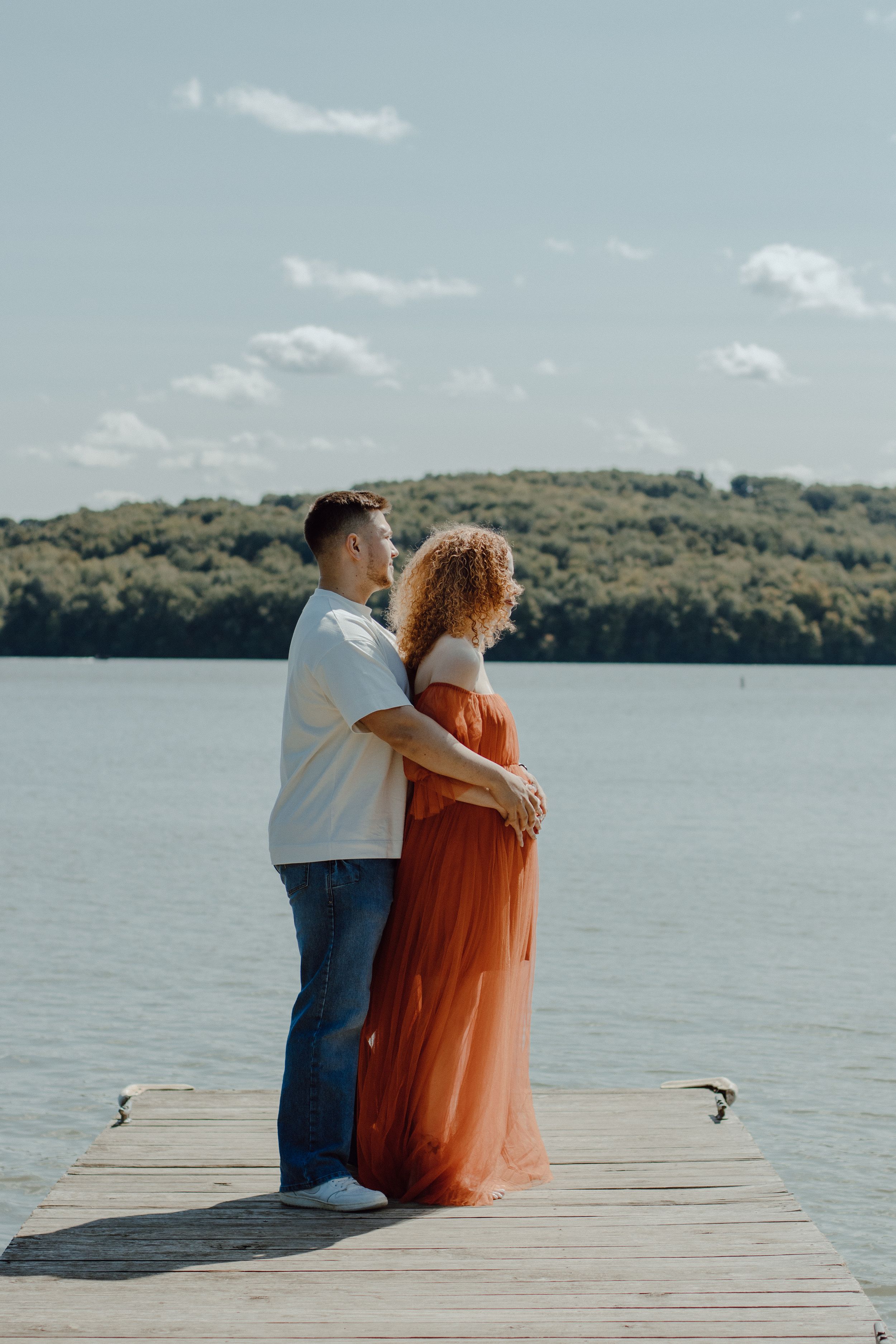 Couple in white shirt and orange dress embracing on lakeside dock.