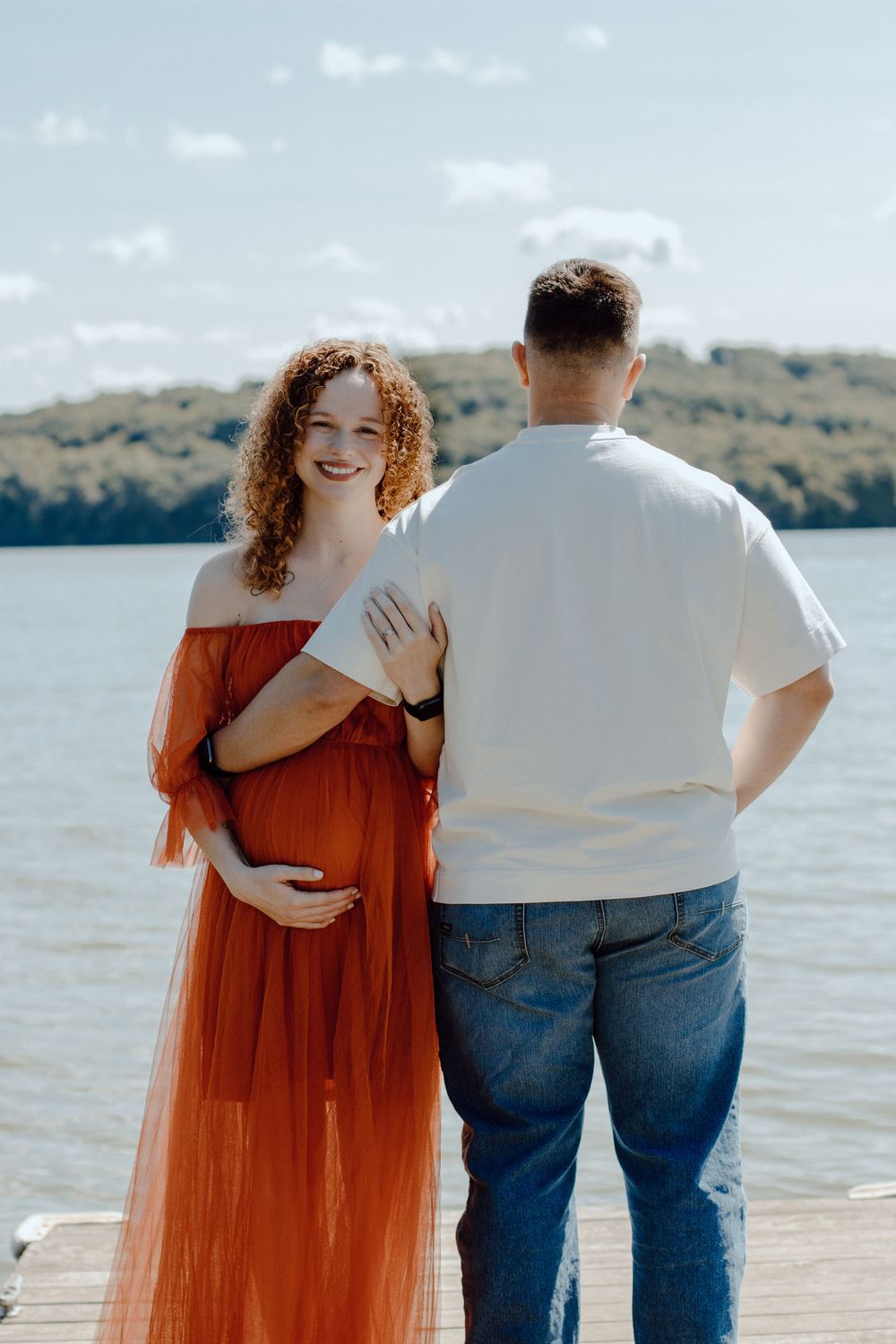Couple's maternity photos on wooden dock with rust-colored dress and casual attire.