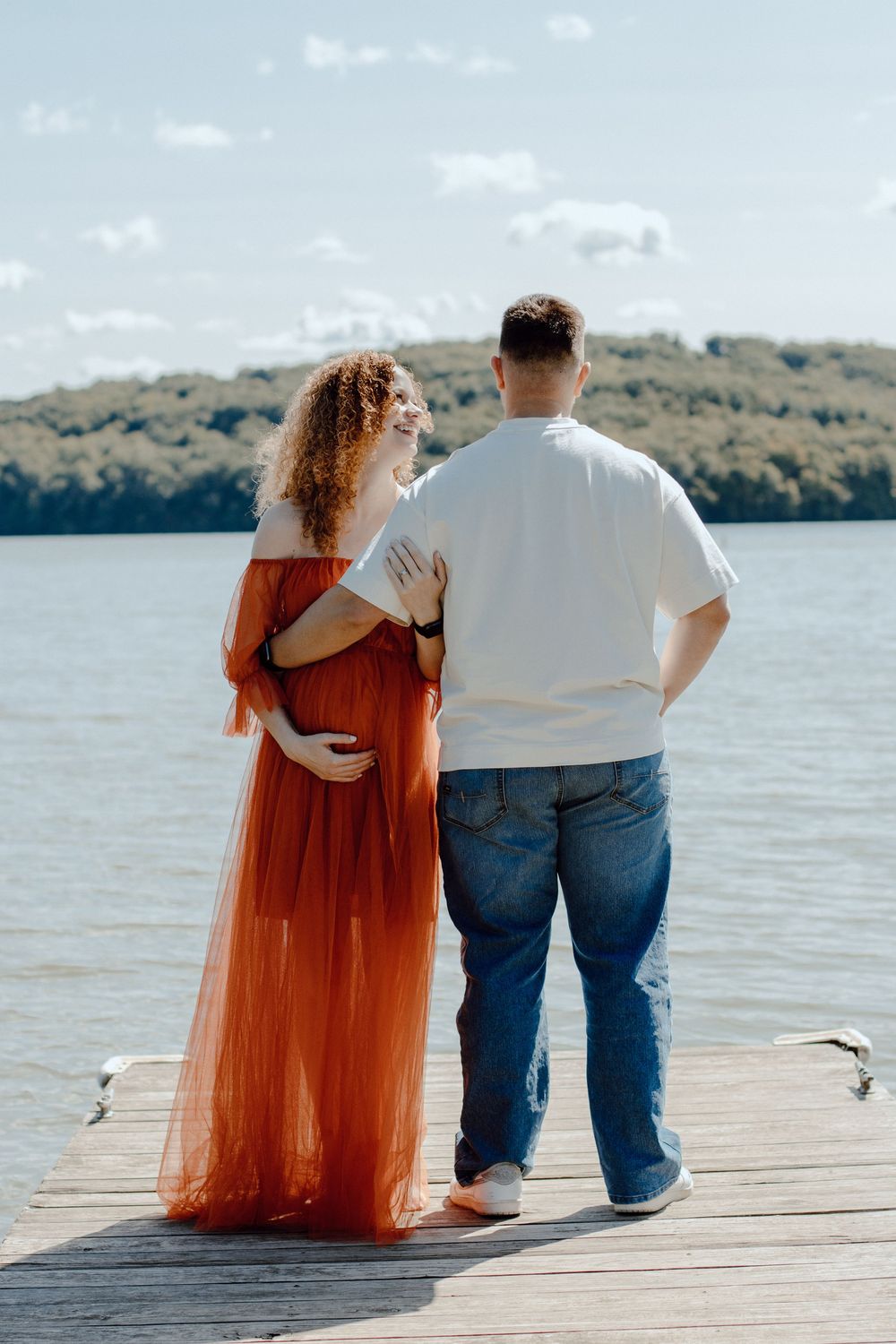 Couple standing on wooden dock by lake with woman in flowing orange dress and man in white shirt.