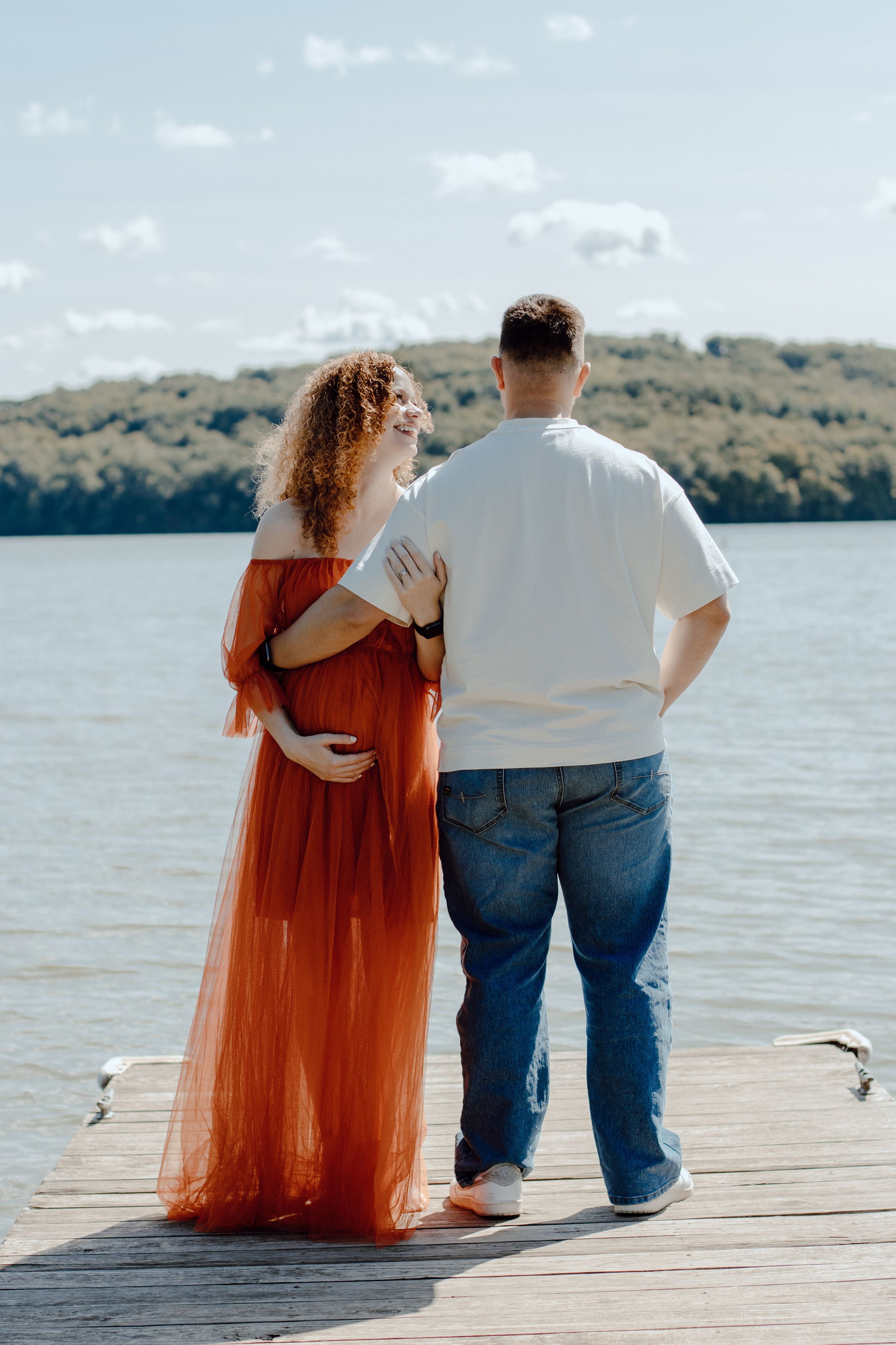 Couple standing on wooden dock by lake with woman in flowing orange dress and man in white shirt.
