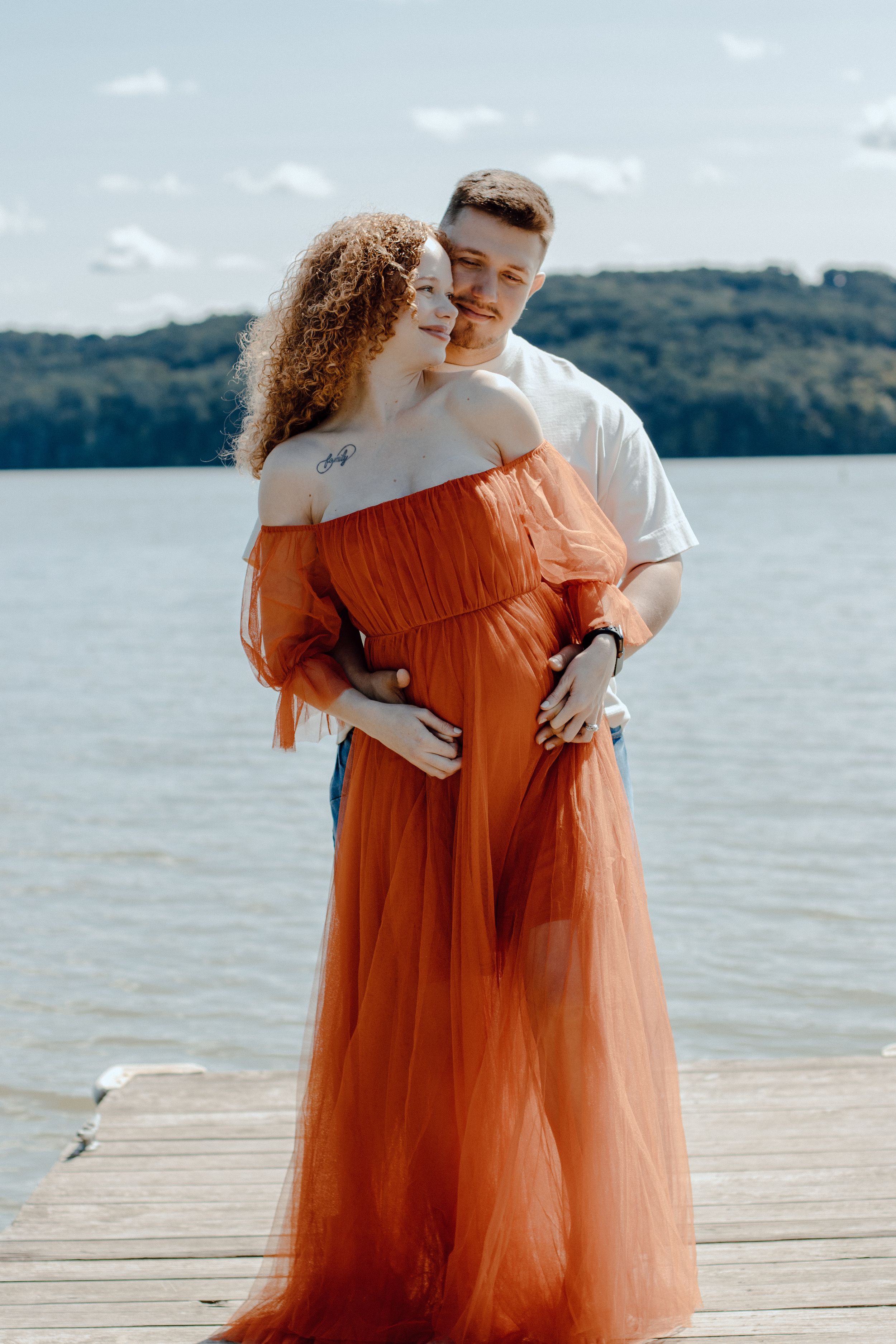Romantic couple embracing on dock with woman wearing orange maxi dress against lakeside backdrop.