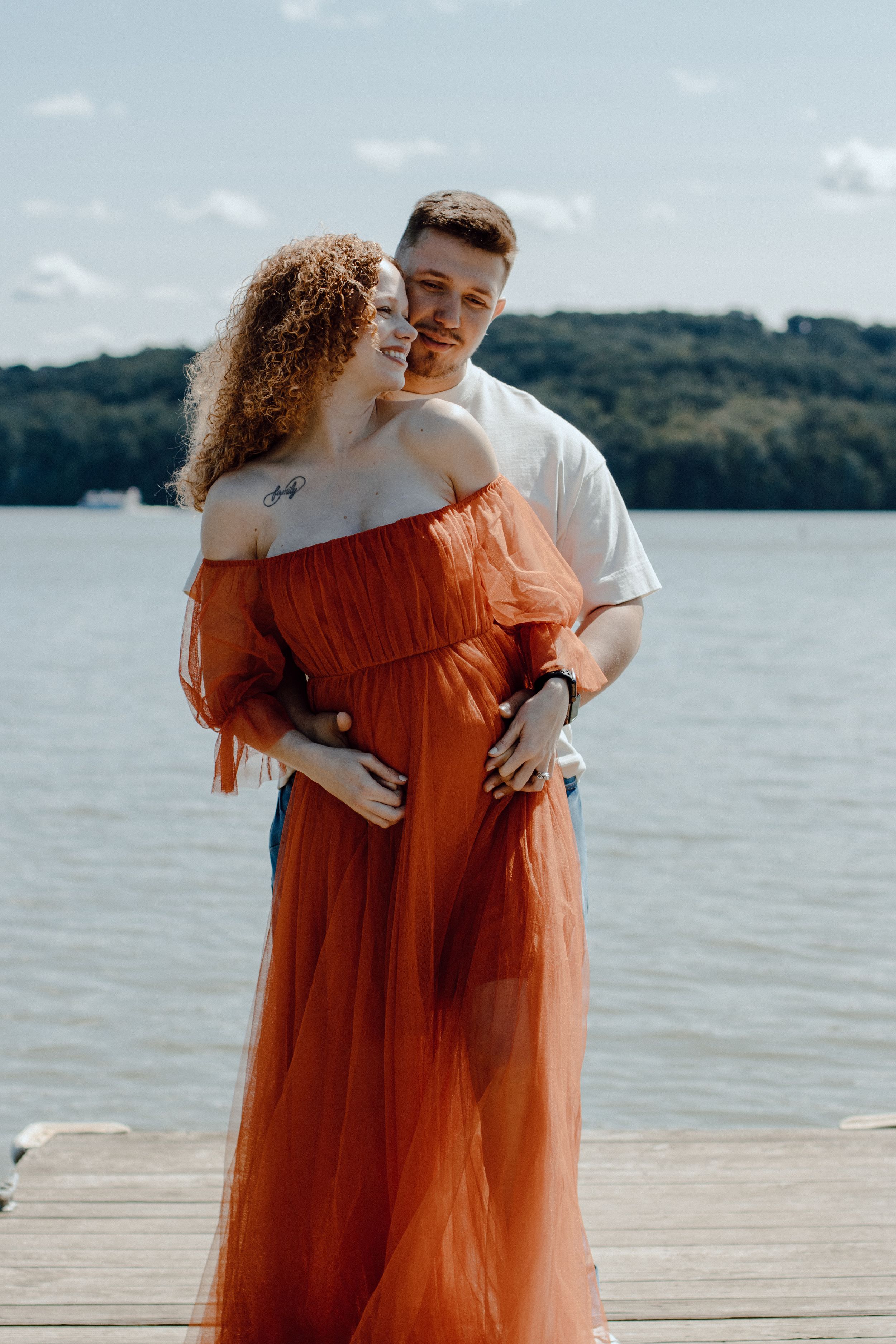 Intimate moment between couple on lakeside dock with orange dress flowing in breeze.