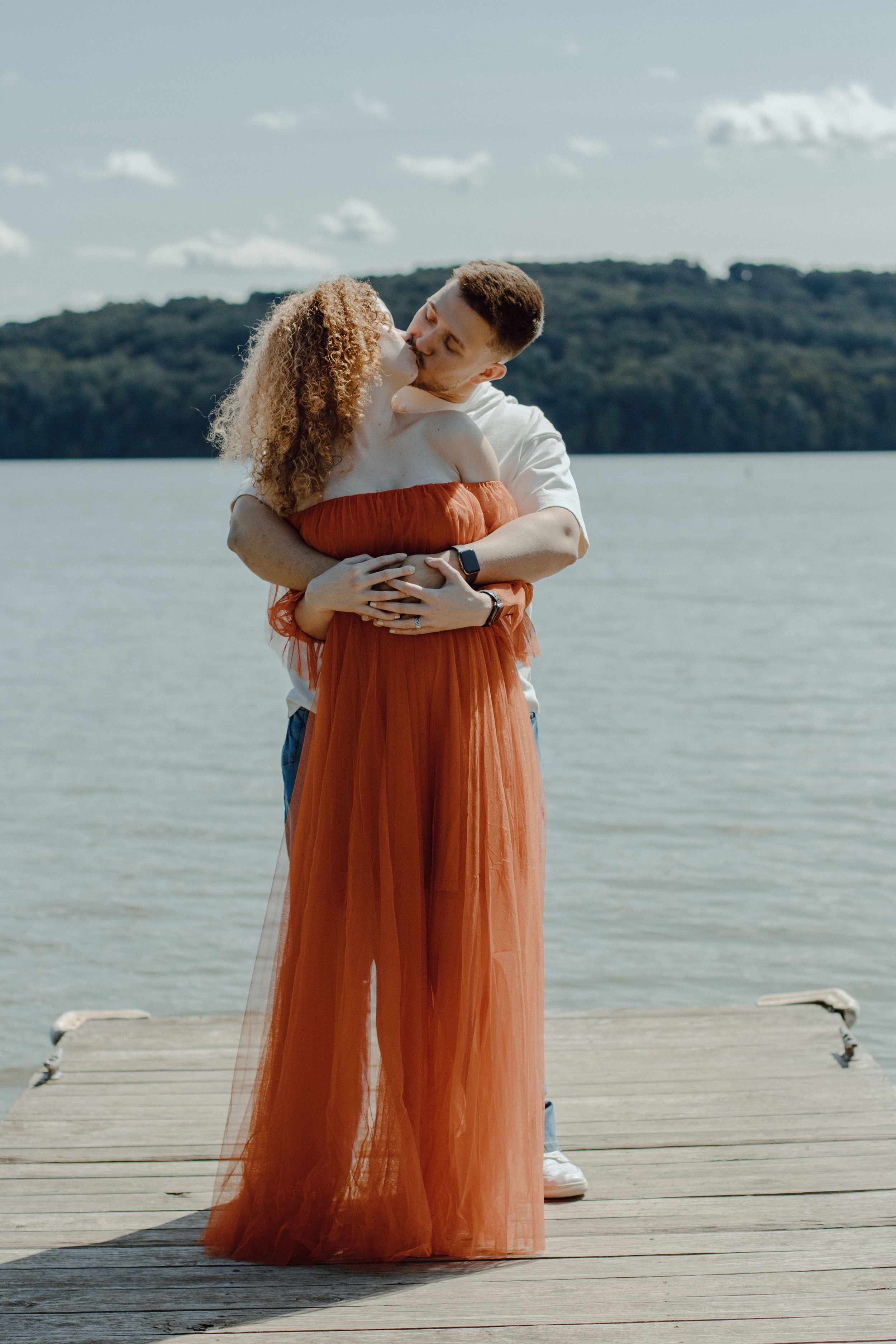 Tender embrace between couple on wooden pier with scenic lake view in background.