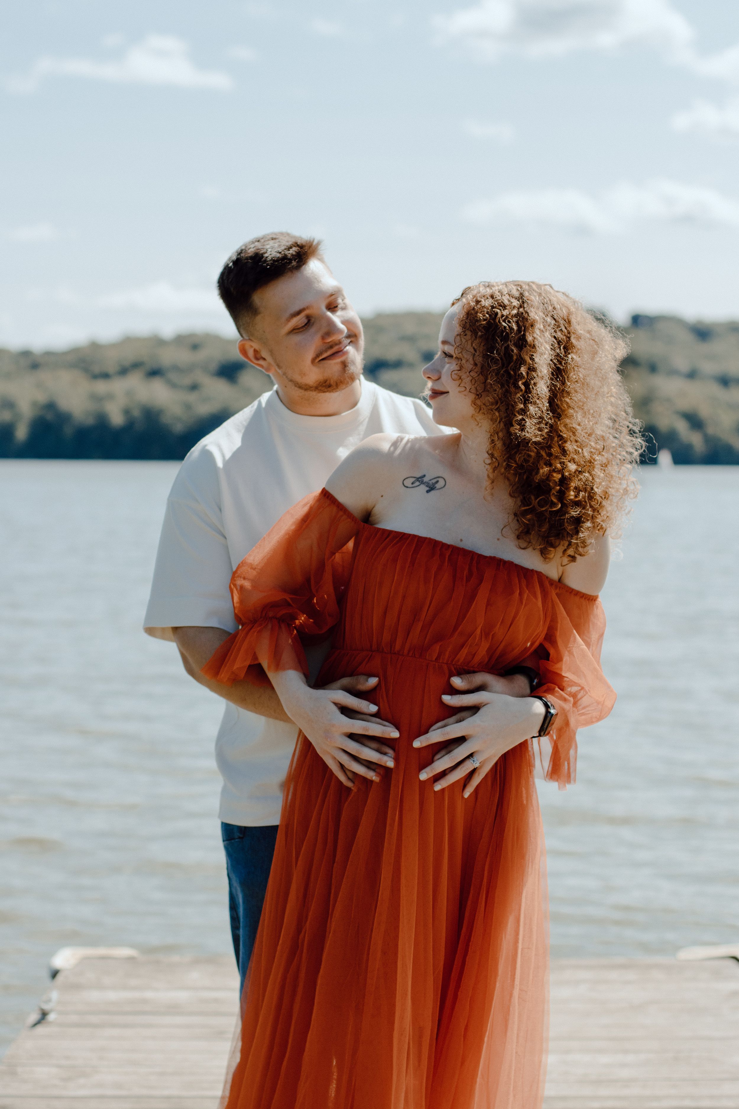 Couple sharing loving moment on dock with woman in orange off-shoulder dress.