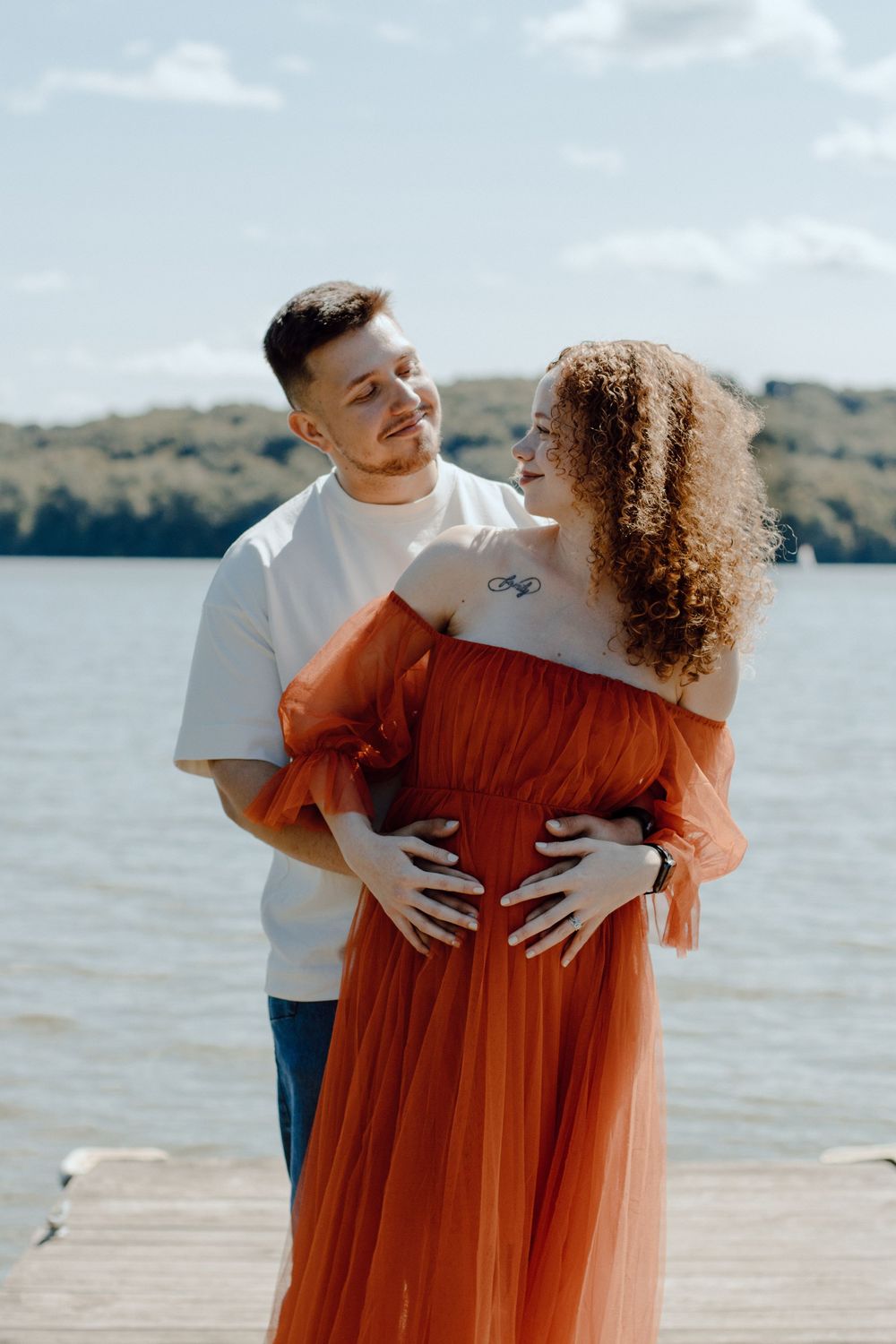 Couple sharing loving moment on dock with woman in orange off-shoulder dress.