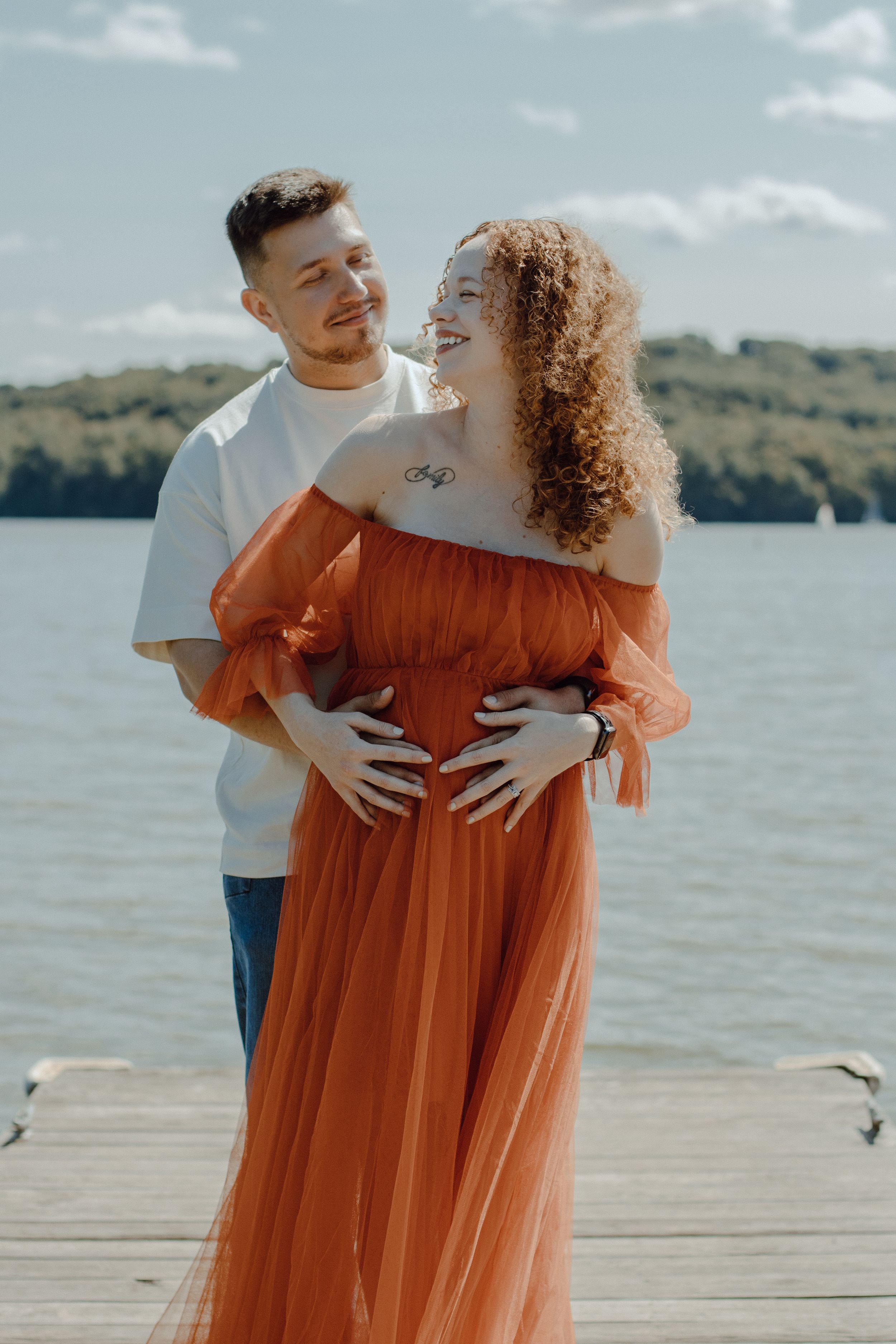 Happy couple embracing on lakeside dock with woman in flowing orange gown.