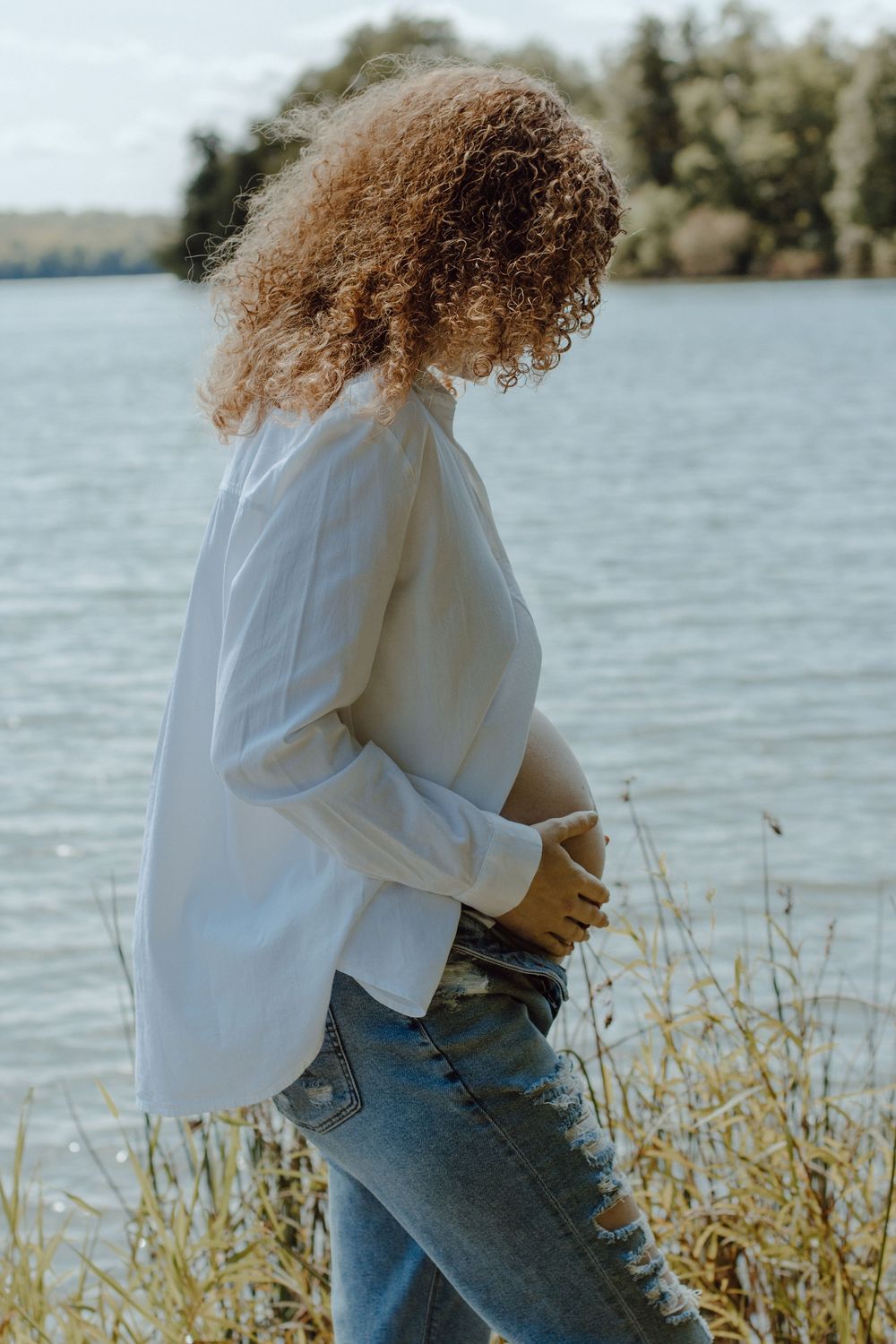 Figure in white sweater and jeans poses near lake with sunlit water in background.