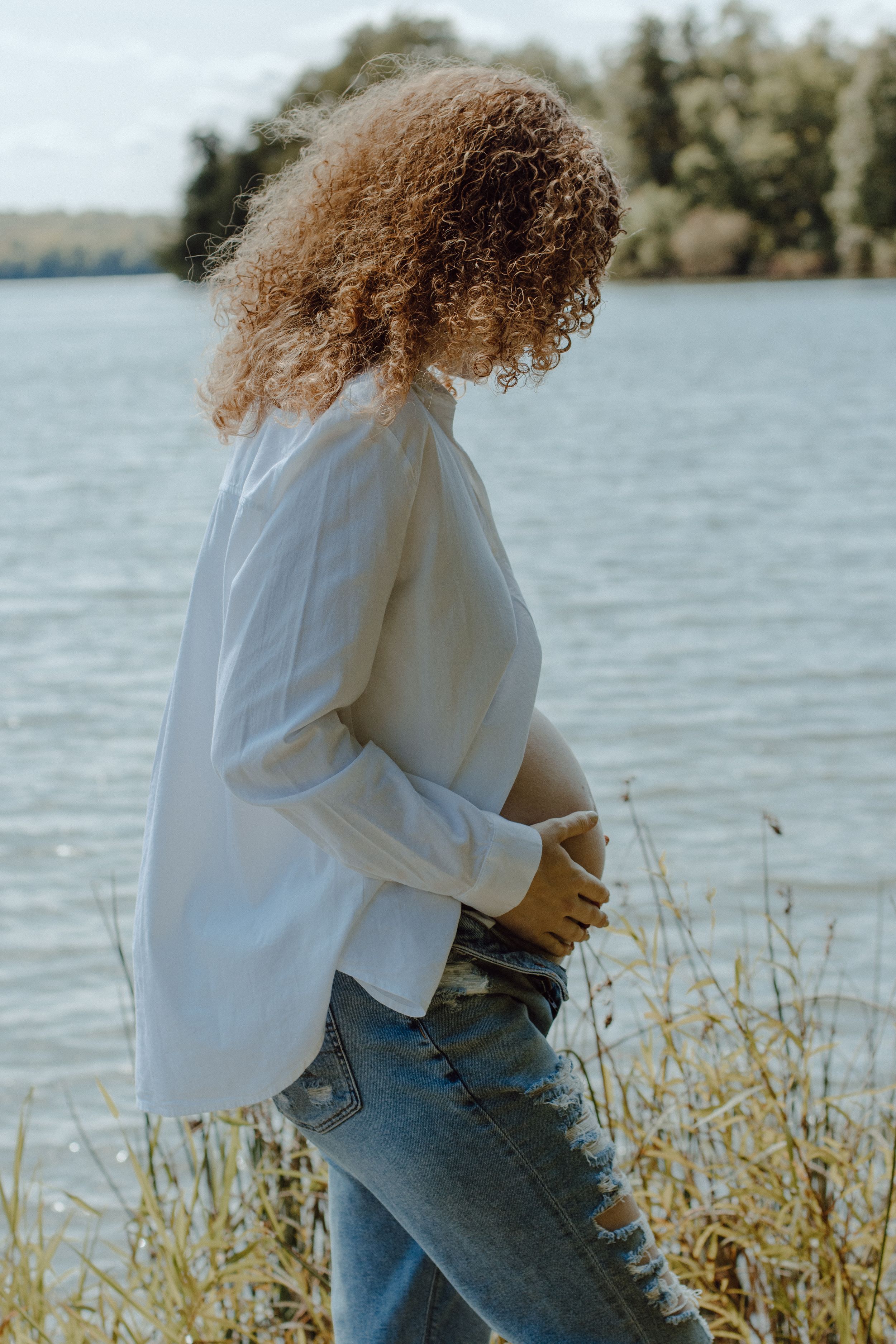 Figure in white sweater and jeans poses near lake with sunlit water in background.