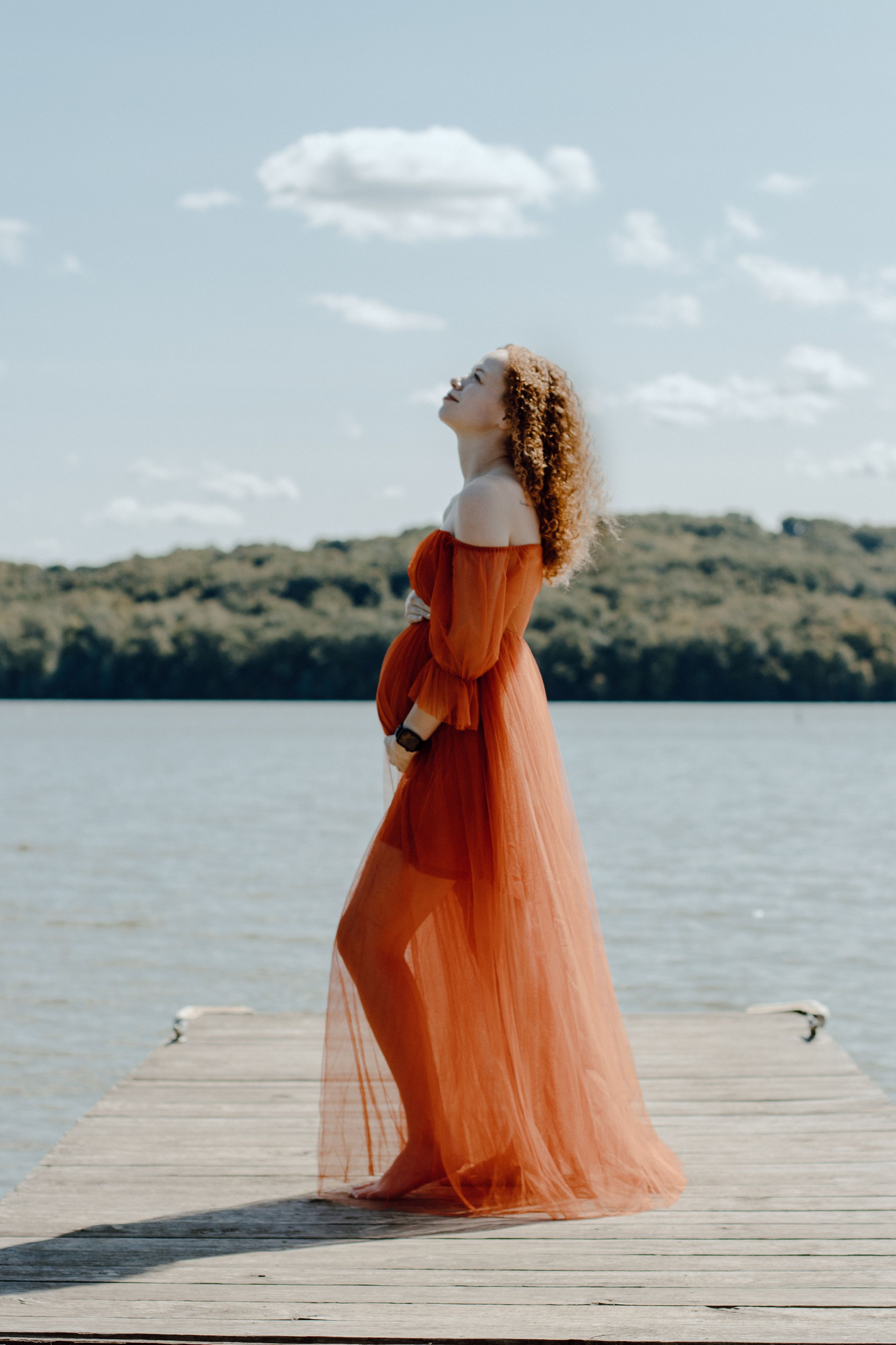 Woman in orange tulle dress standing alone on dock looking up at sky.