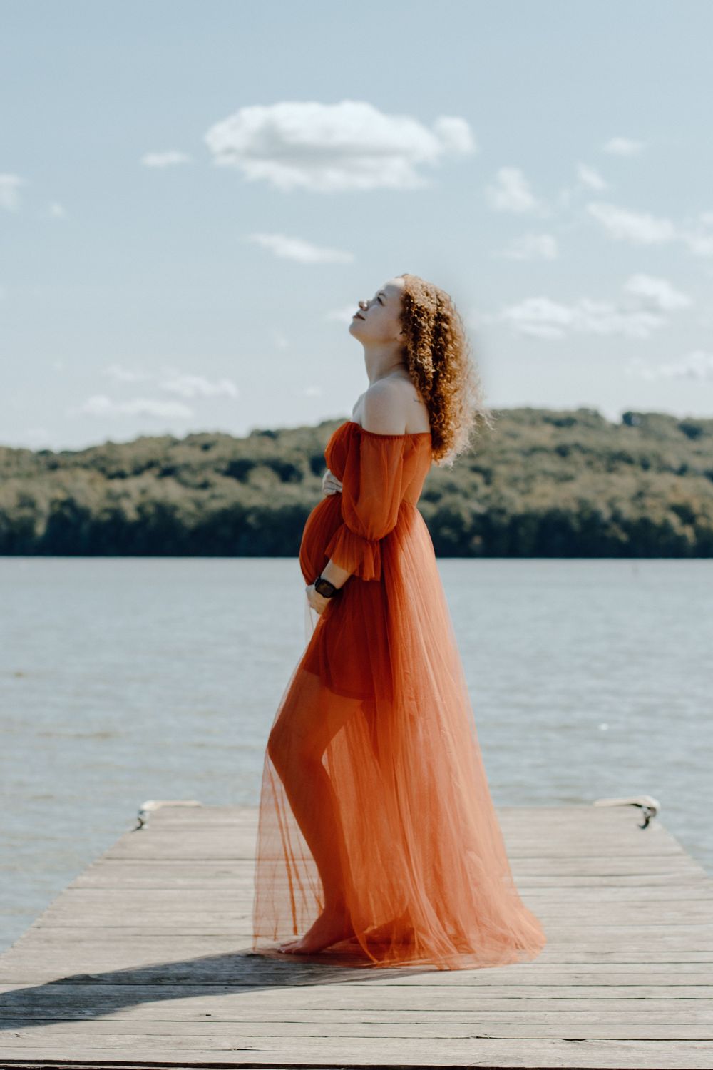 Woman in orange tulle dress standing alone on dock looking up at sky.